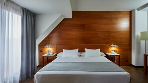 Una habitación de hotel moderna con una cama grande, cabecera de madera, dos mesitas de noche con lámparas y vistas al exterior.