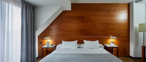 A modern hotel room with a large bed, wooden headboard, two bedside tables with lamps, and a view of the outdoors.