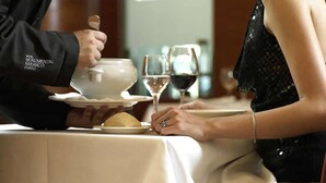 Una persona sirve un plato blanco a otra persona sentada a una mesa, con una copa de vino.