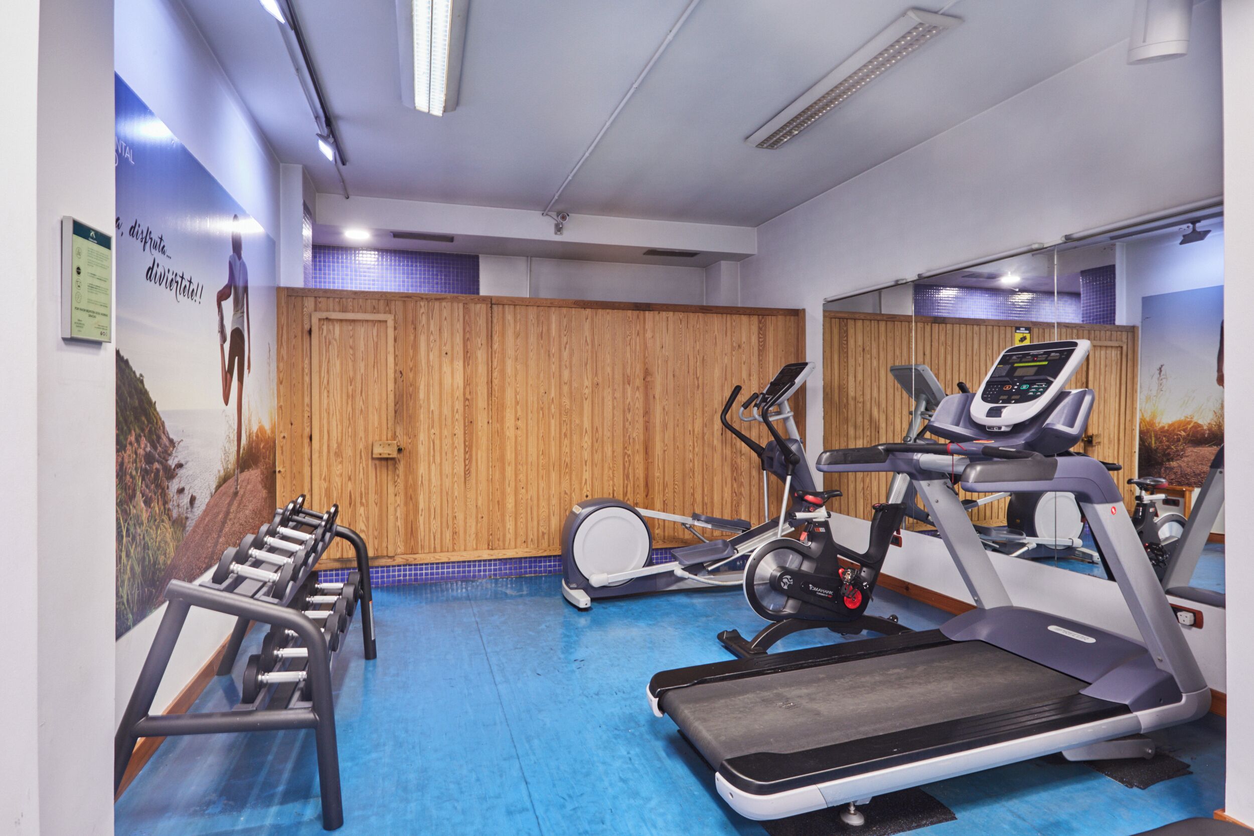 Une salle de sport équipée de matériel d’exercice, avec des murs recouverts de panneaux en bois et un sol bleu.