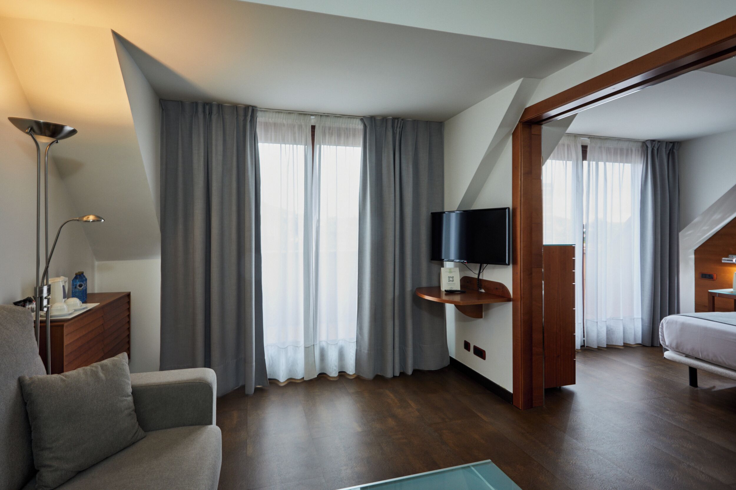 A modern hotel room with a sofa, a flat-screen TV, a wooden side table, and a large window with curtains.