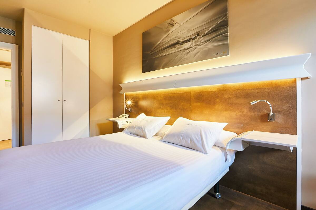 Une chambre d’hôtel avec un grand lit, une table de chevet avec une lampe, une étagère fixée au mur et une porte située à gauche.