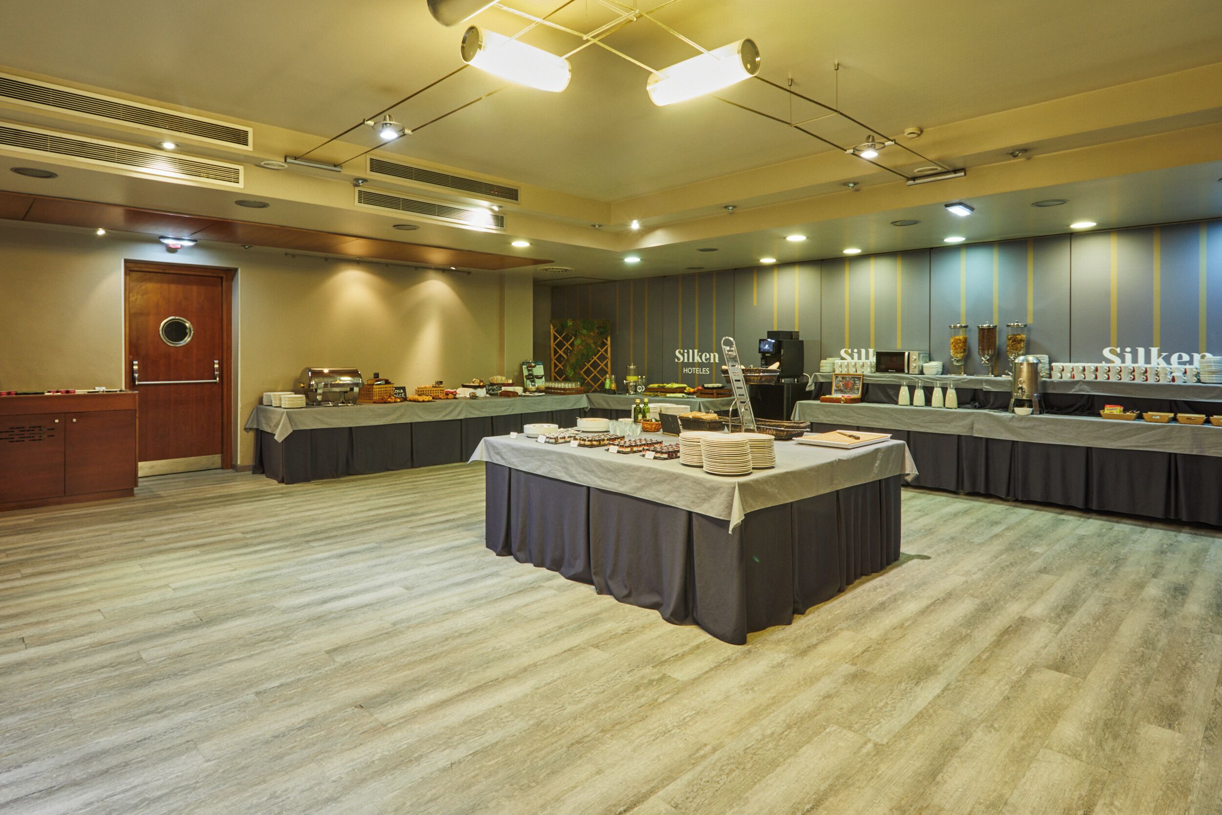A spacious banquet hall with a long table covered in a tablecloth, surrounded by buffet stations with various food items and a coffee machine.