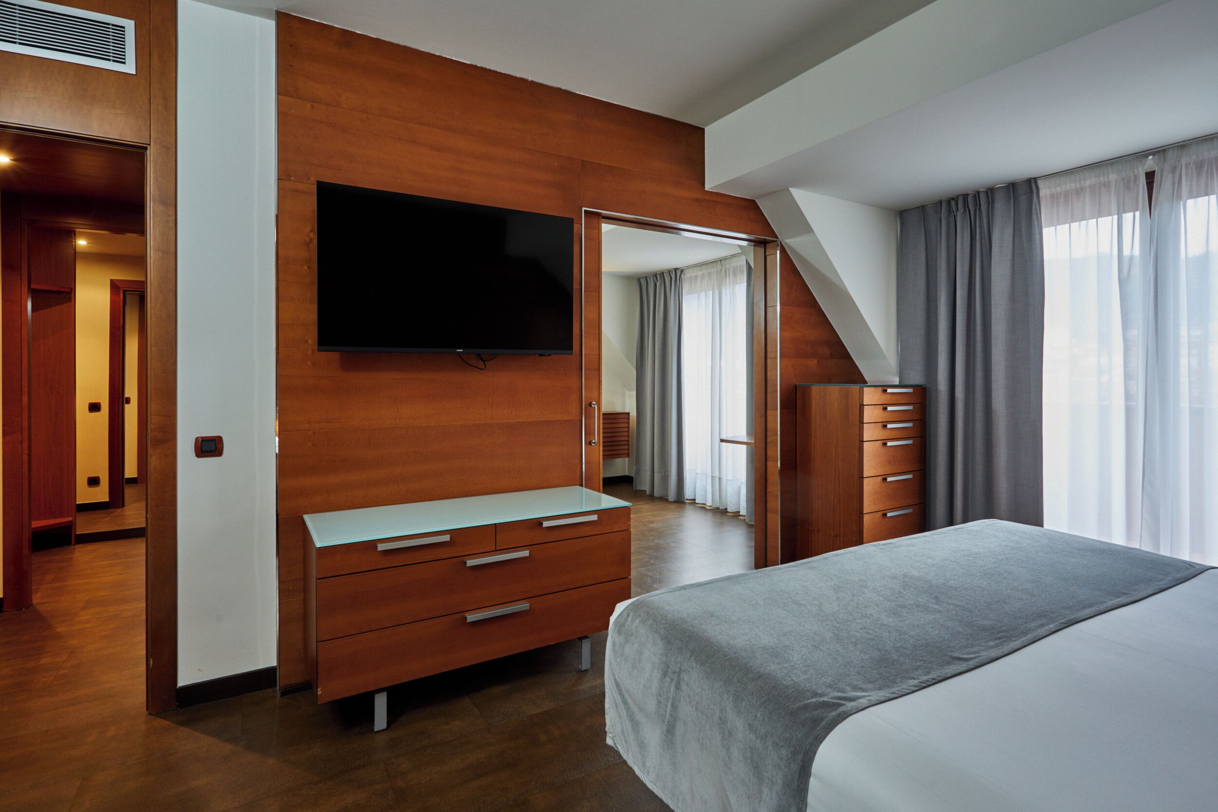 A modern hotel room with a large bed, a flat-screen TV mounted on the wall, a wooden dresser, and a mirror.