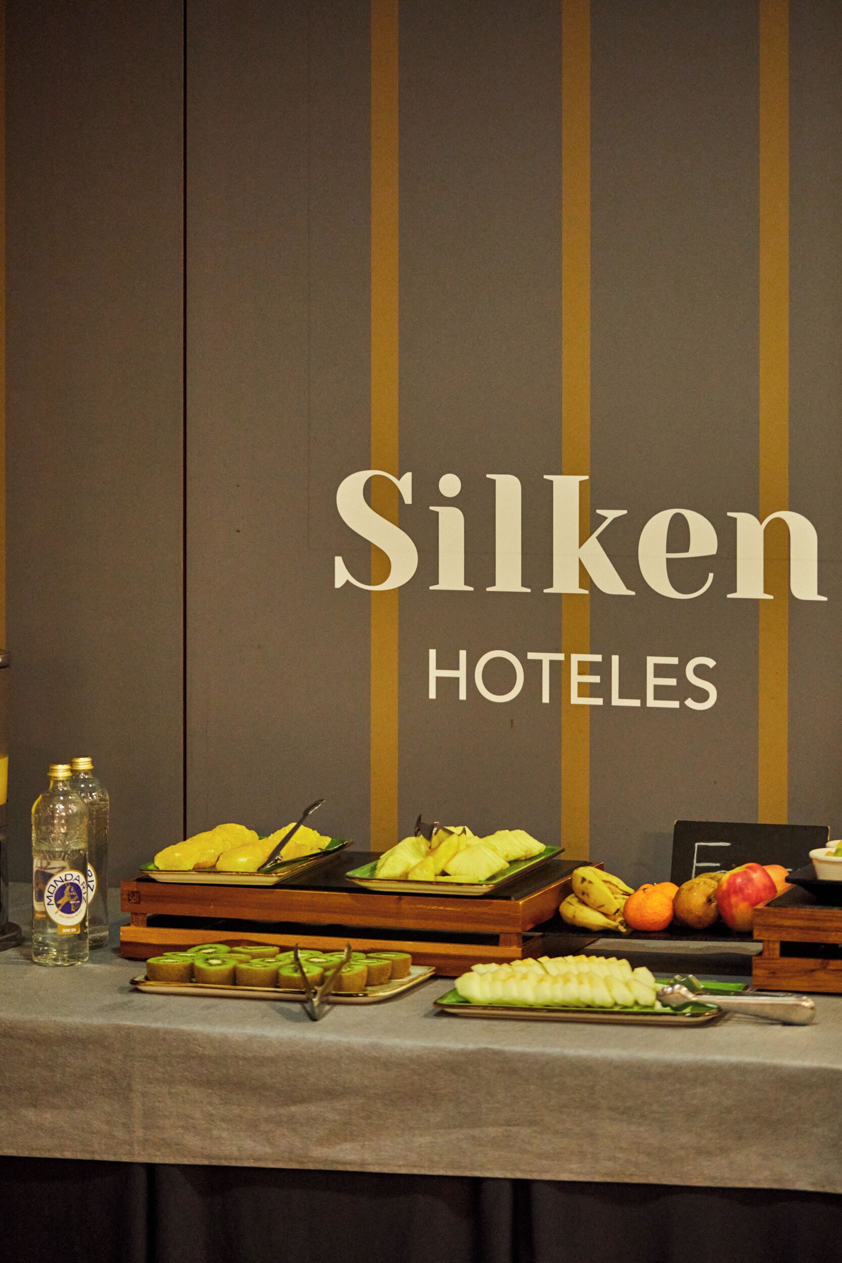 A buffet table with various fruits and a bottle of water, set against a wall with the text "Silken HOTELES".