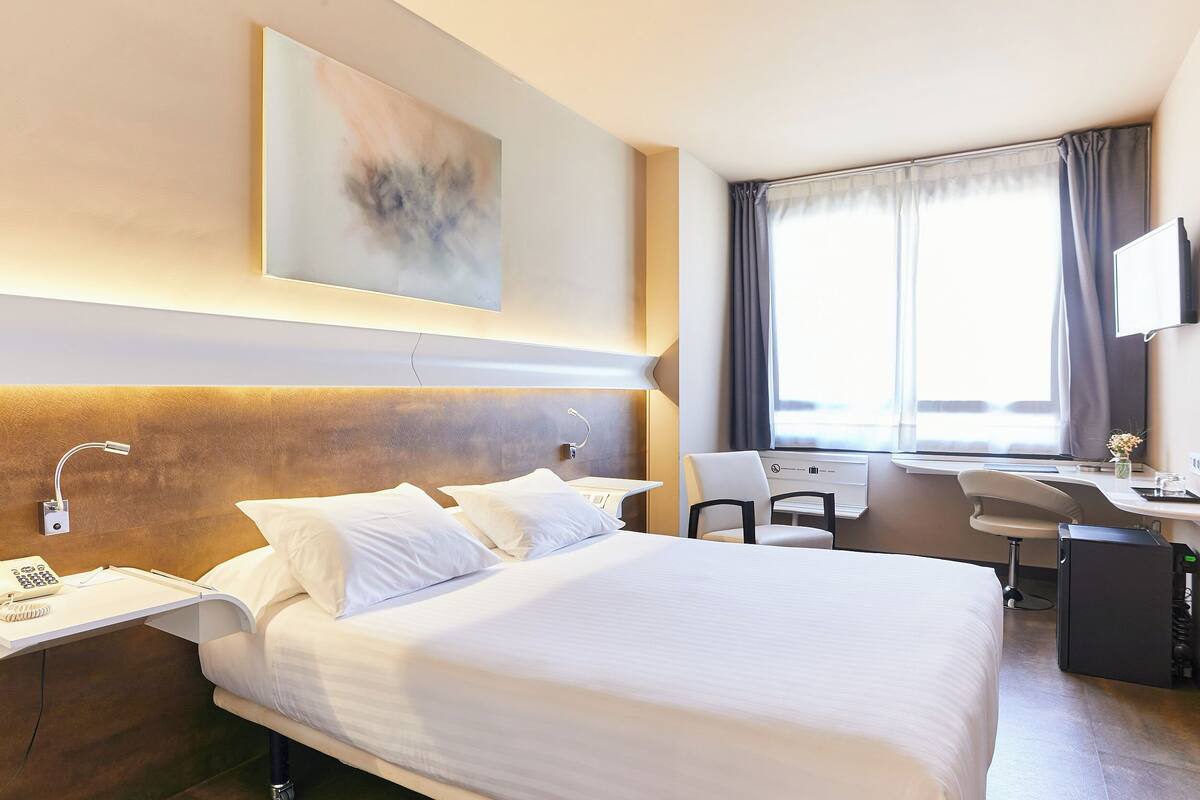 Une chambre d’hôtel avec un grand lit, un bureau, une chaise, une lampe de bureau, un téléphone et un tableau au mur.