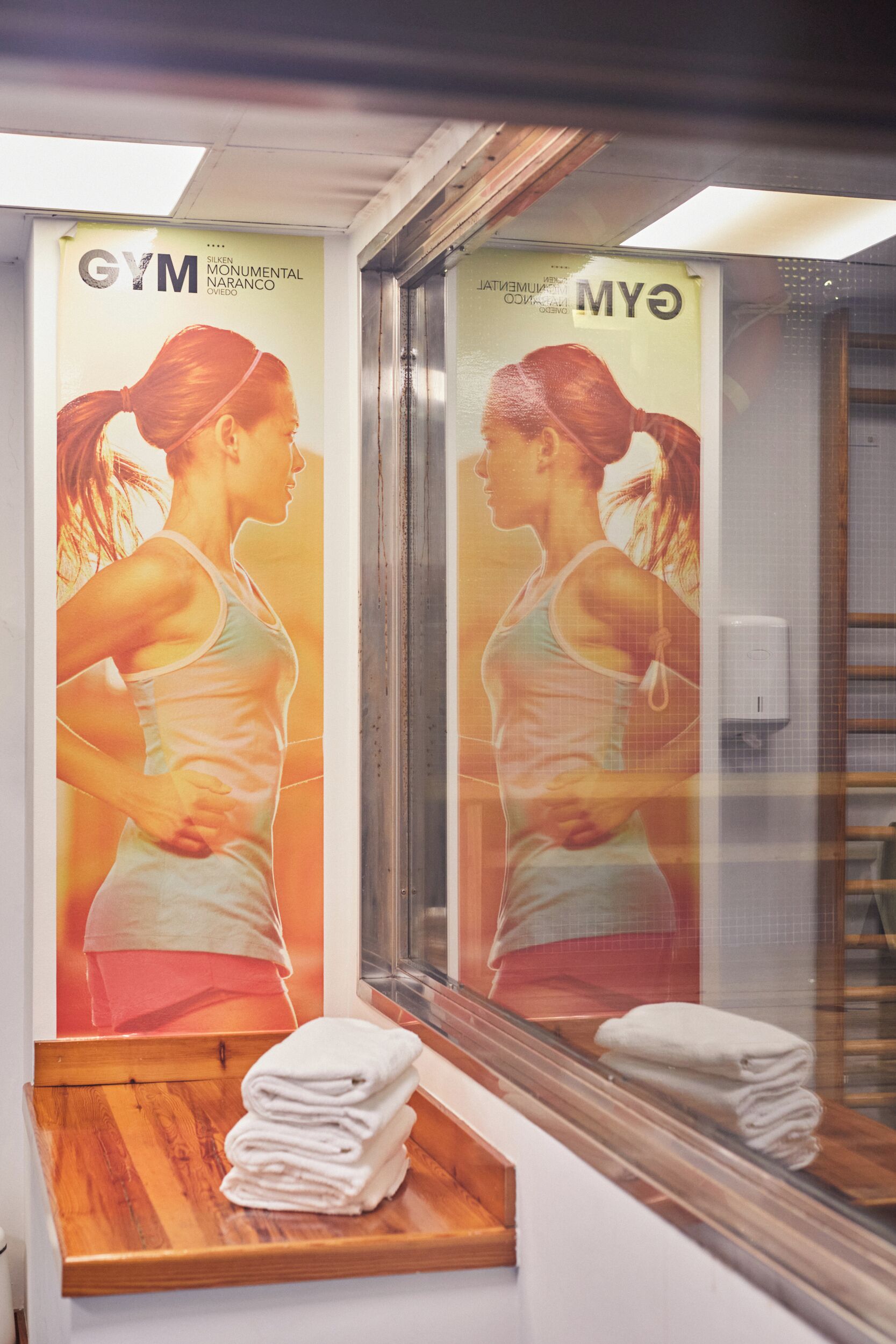 Un gimnasio con un espejo grande, un banco de madera con toallas y un cartel de una mujer ejercitándose.