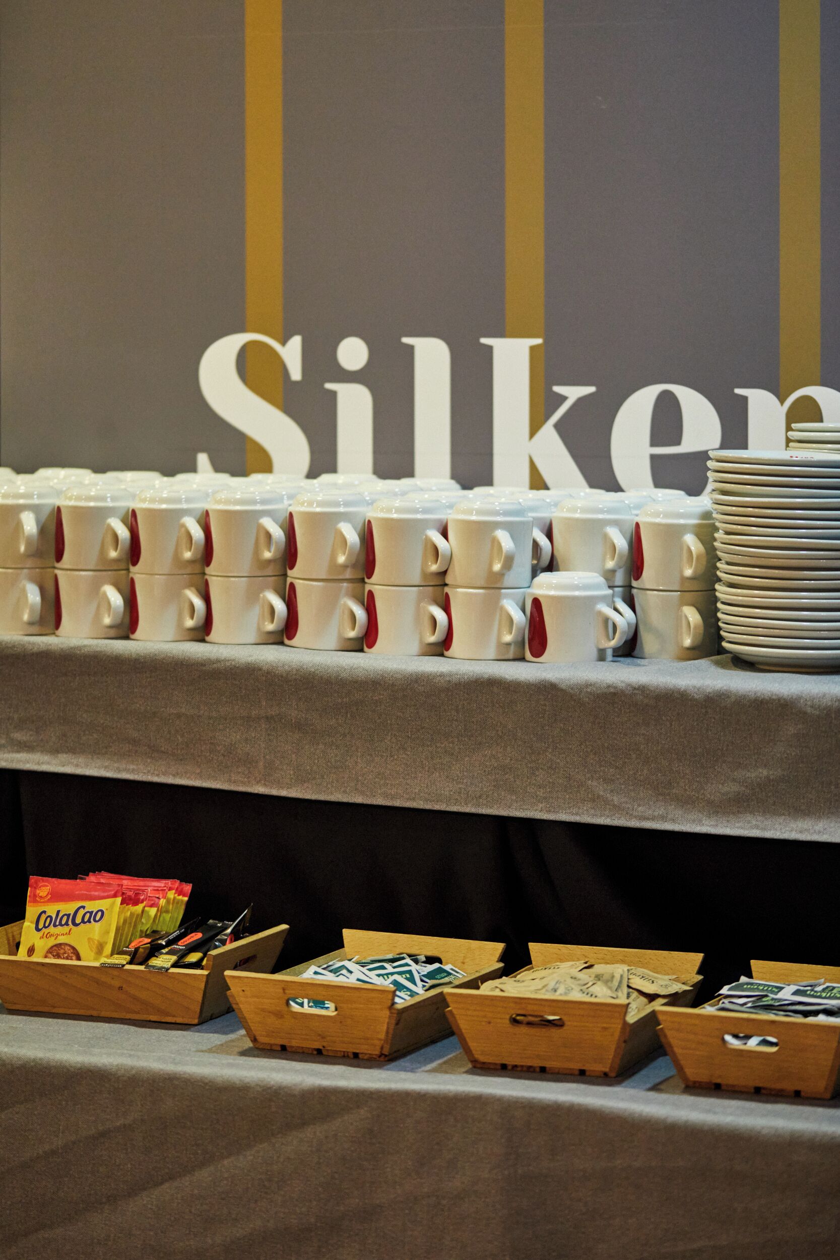 A table with stacked white mugs and plates, and wooden boxes containing snacks.