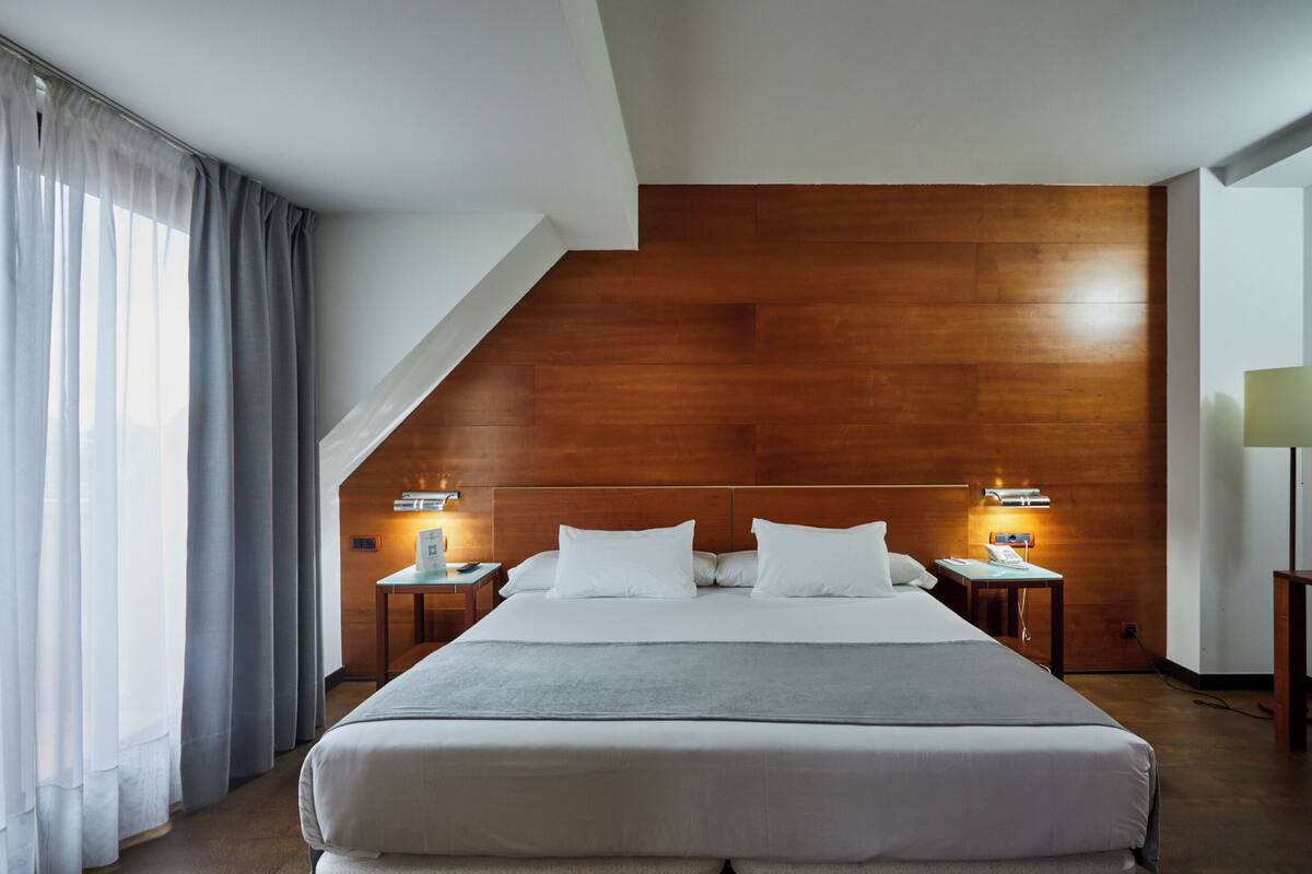 Une chambre d’hôtel moderne avec un grand lit, une tête de lit en bois, deux tables de chevet avec des lampes et une vue sur l’extérieur par une fenêtre ornée de rideaux légers.