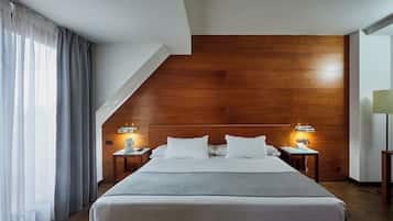 A modern hotel room with a large bed, wooden headboard, two bedside tables with lamps, and a view of the outdoors through a window with sheer curtains.
