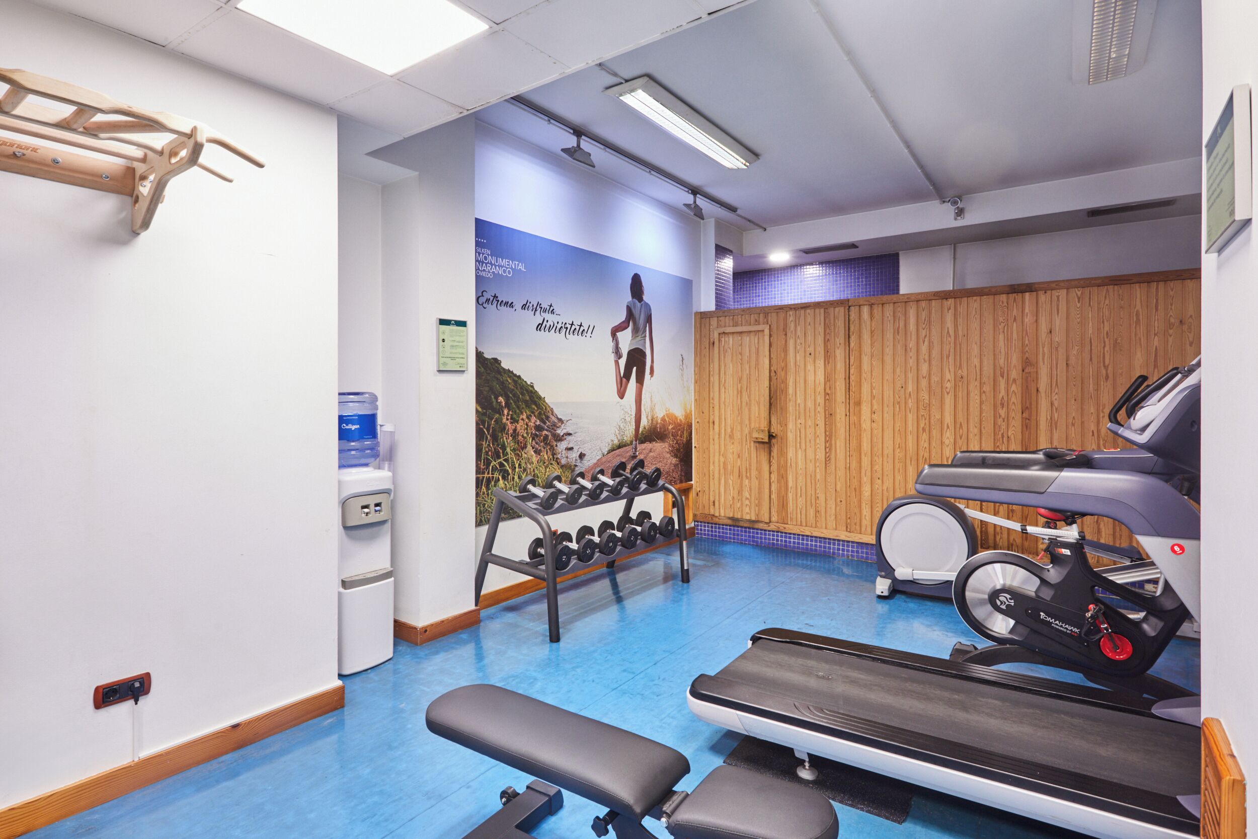 Un gimnasio con equipamiento de ejercicio, dispensador de agua y un mural en la pared.