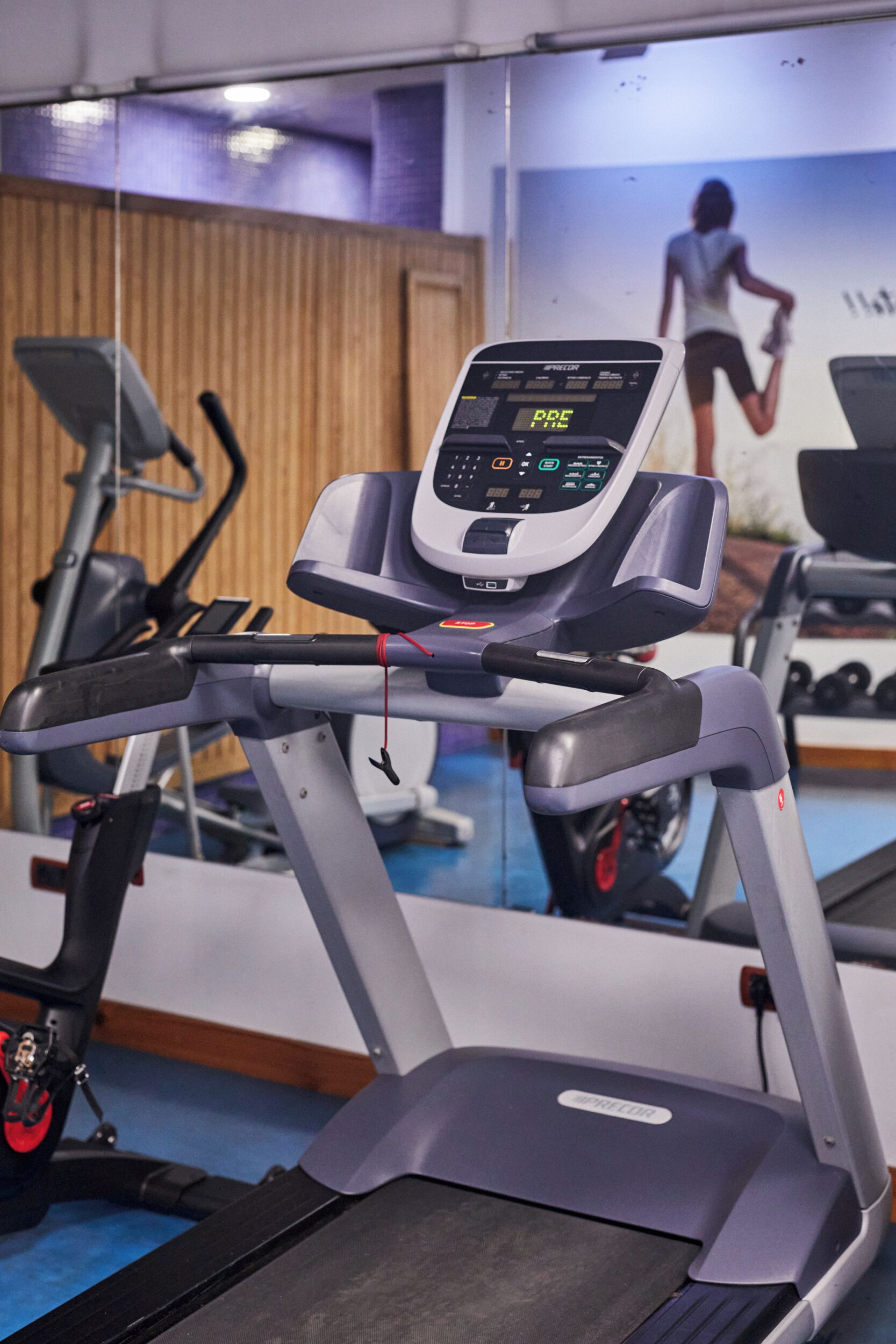 A treadmill with a digital display, a mirror reflecting gym equipment, and a wooden wall in the background.