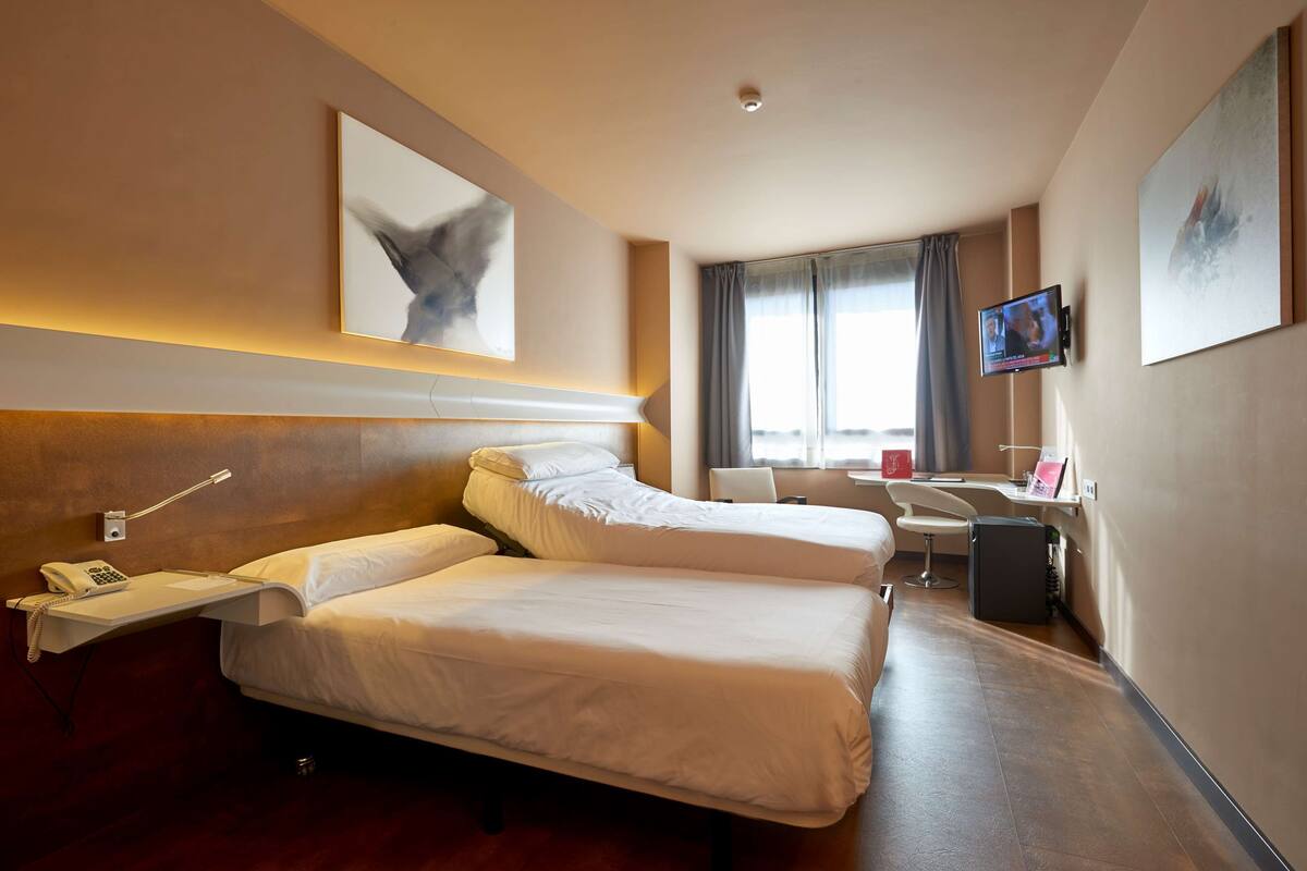 Une chambre d’hôtel avec deux lits, un bureau, une chaise, une télévision et des œuvres d’art accrochées aux murs.