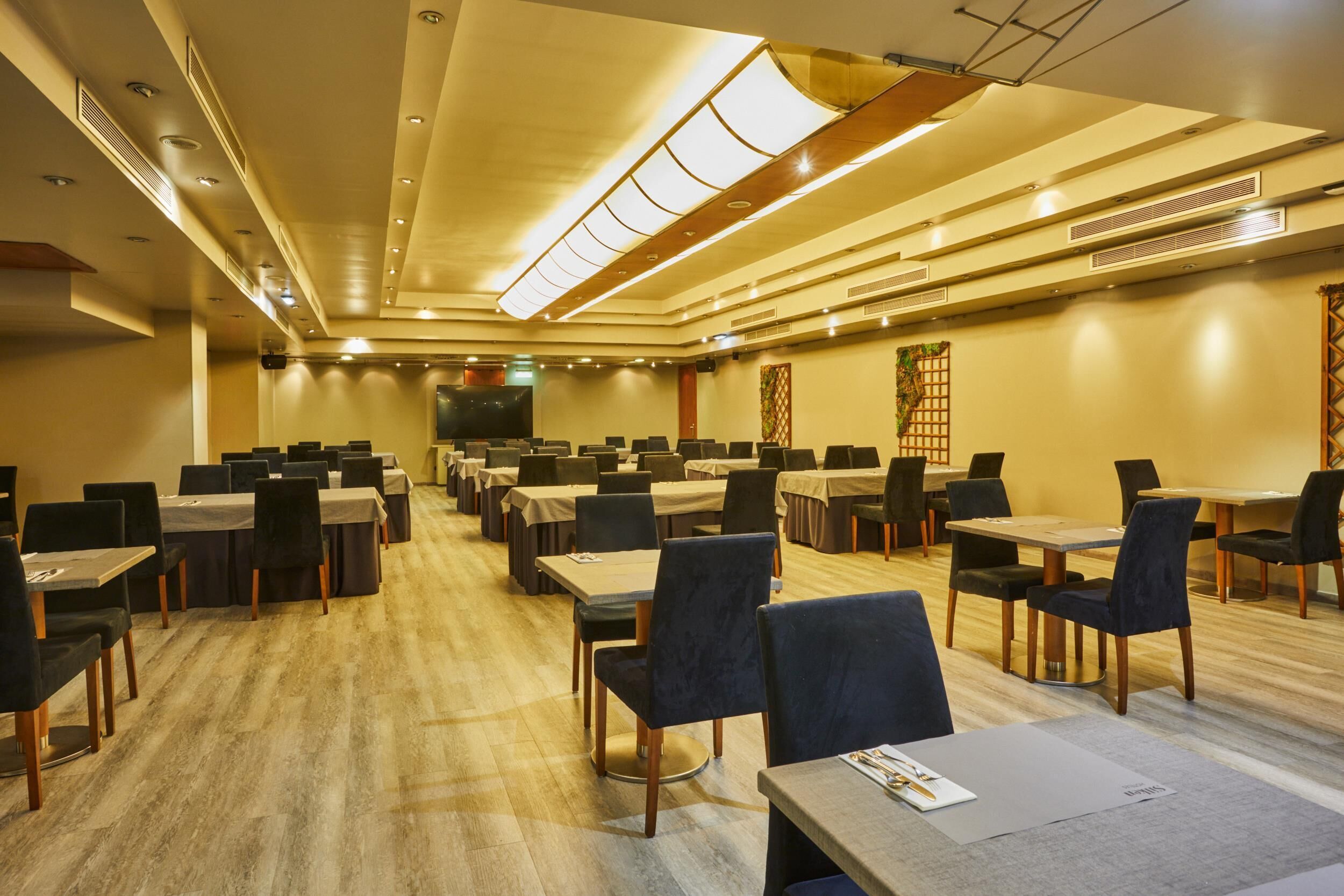 A conference room with multiple tables and chairs, a stage at the front, and a wooden floor.