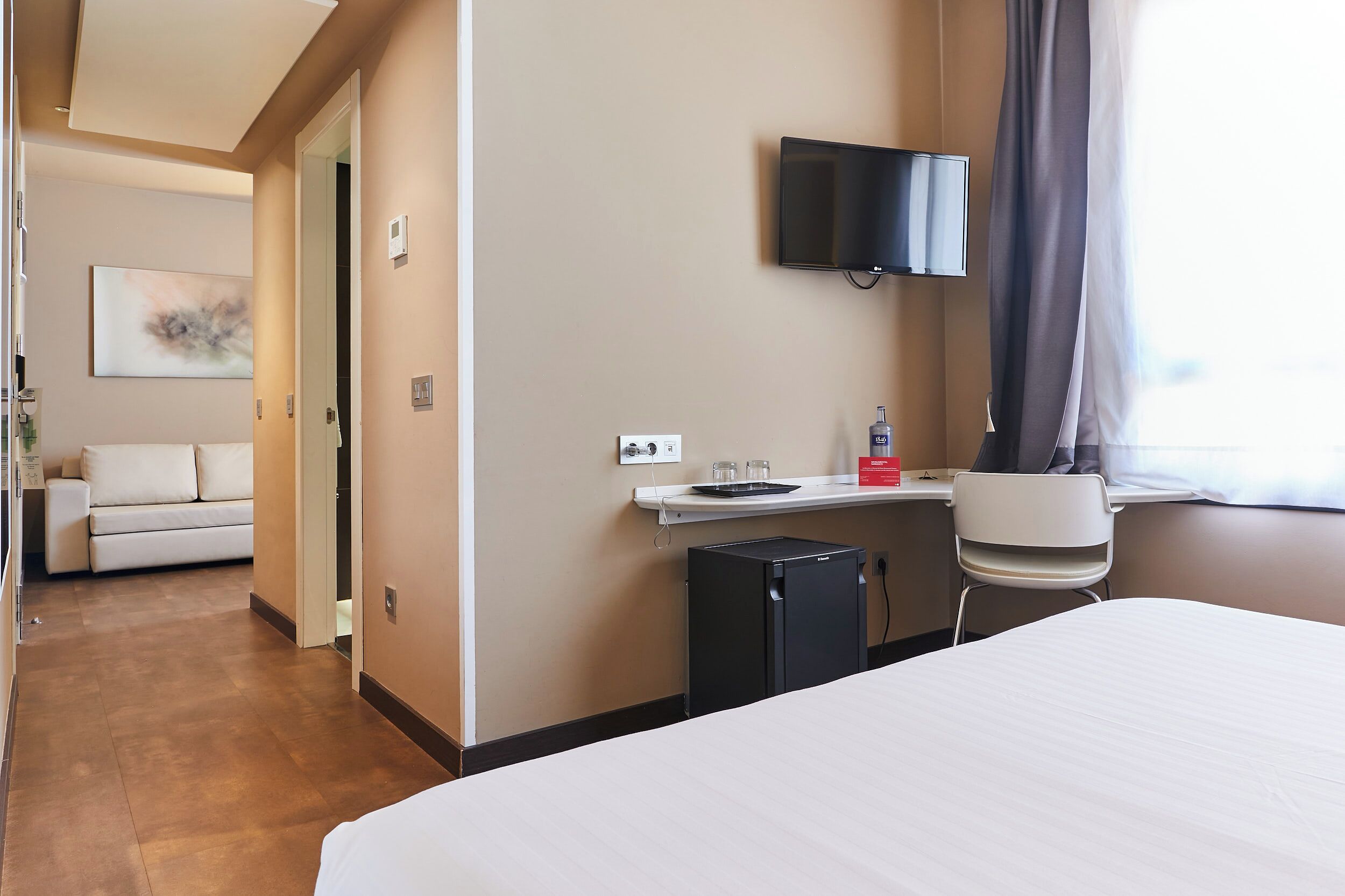 Hotellihuone, jossa on sänky, työpöytä tietokoneella, televisio, pieni jääkaappi ja taustalla oleva sohva.
