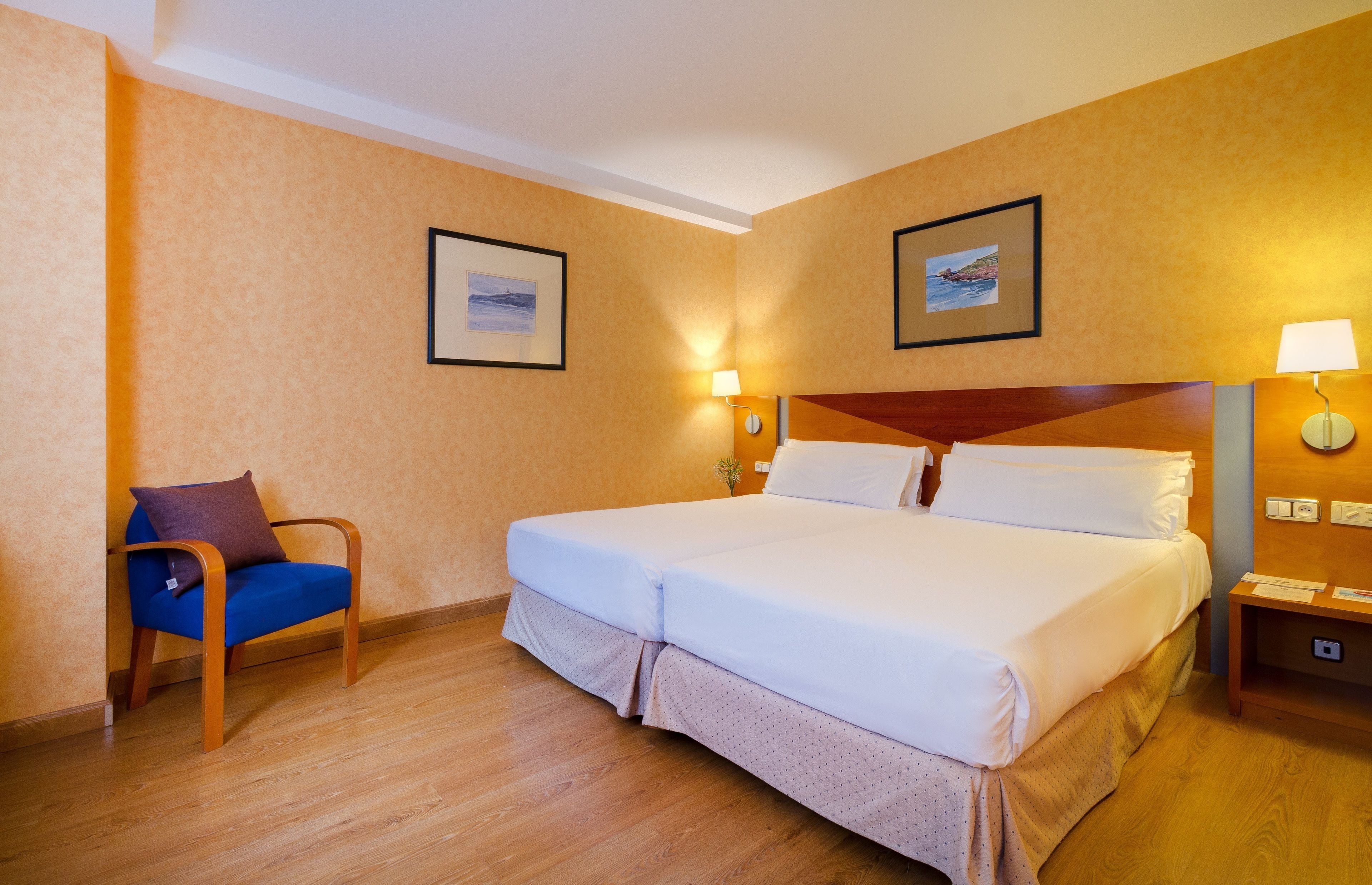 Een hotelkamer met een groot bed, een stoel, twee wandlampen en twee ingekaderde foto’s aan de muur.