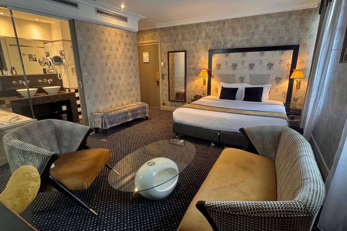 A hotel room with a bed, two armchairs, a coffee table, a desk with a mirror, and a bathroom with a sink and mirror.