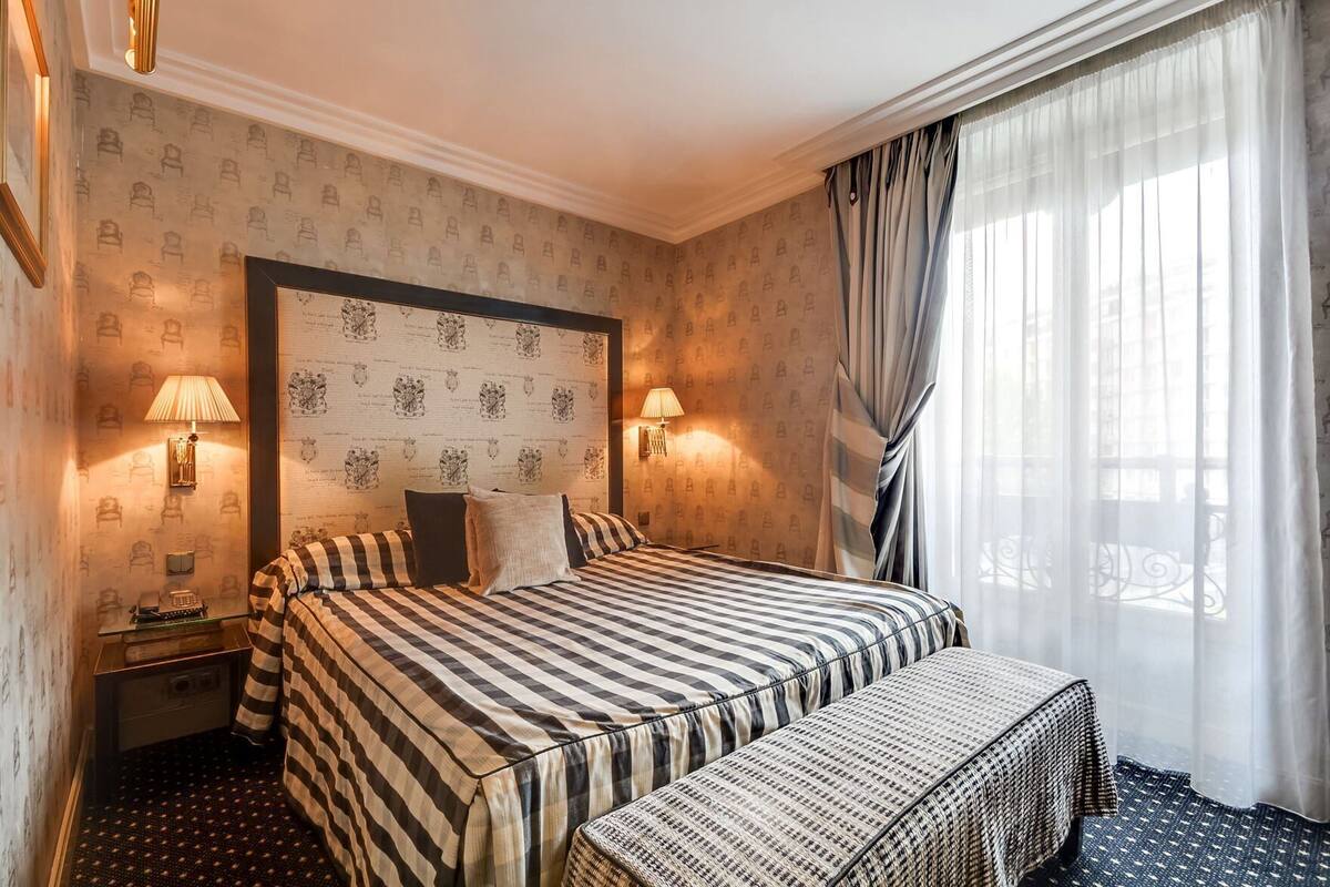 A hotel room with a large bed, two bedside lamps, a headboard with a crest, a small bedside table, and a window with curtains.