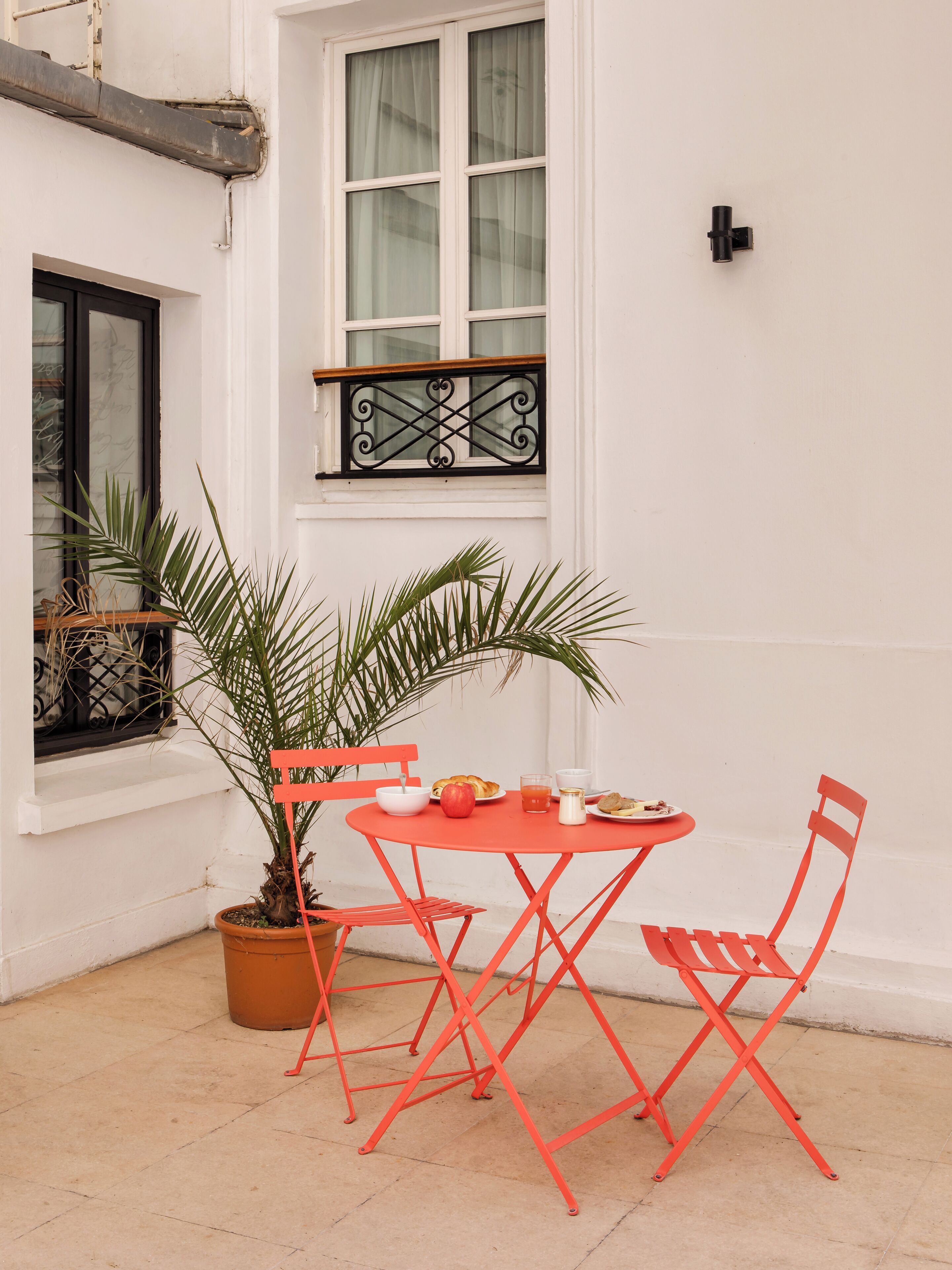 A small outdoor dining area with a table and chairs, a potted plant, and a window with curtains.