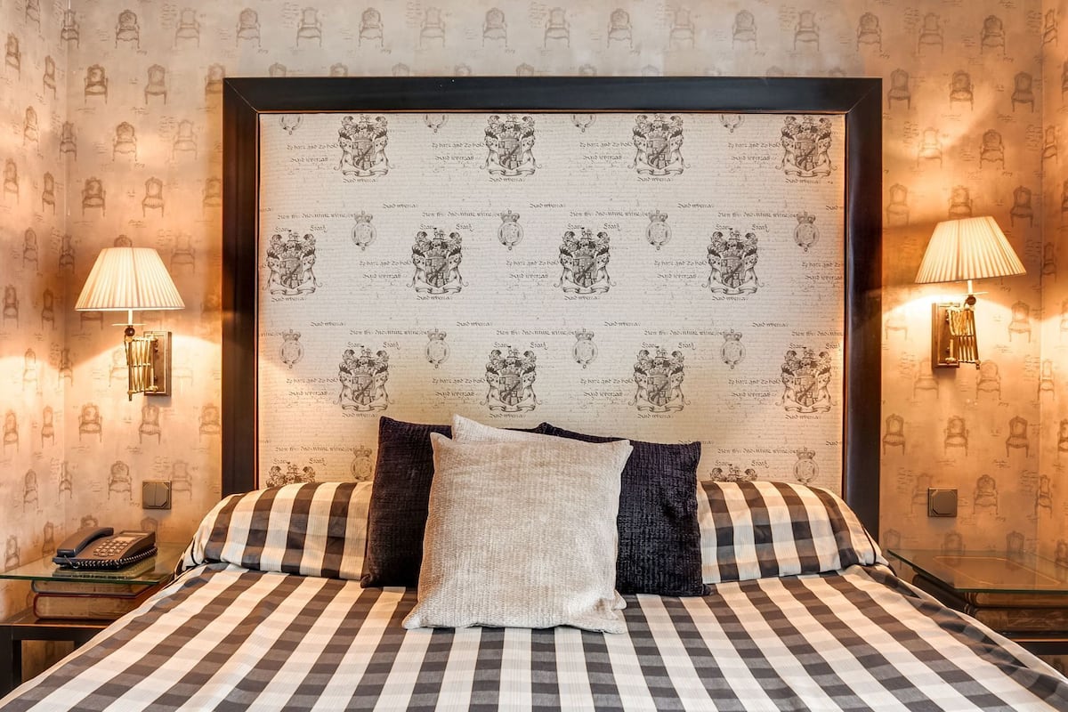 A bed with a checkered bedspread, two pillows, and a headboard with coat of arms. There are two wall-mounted lamps on either side of the bed.