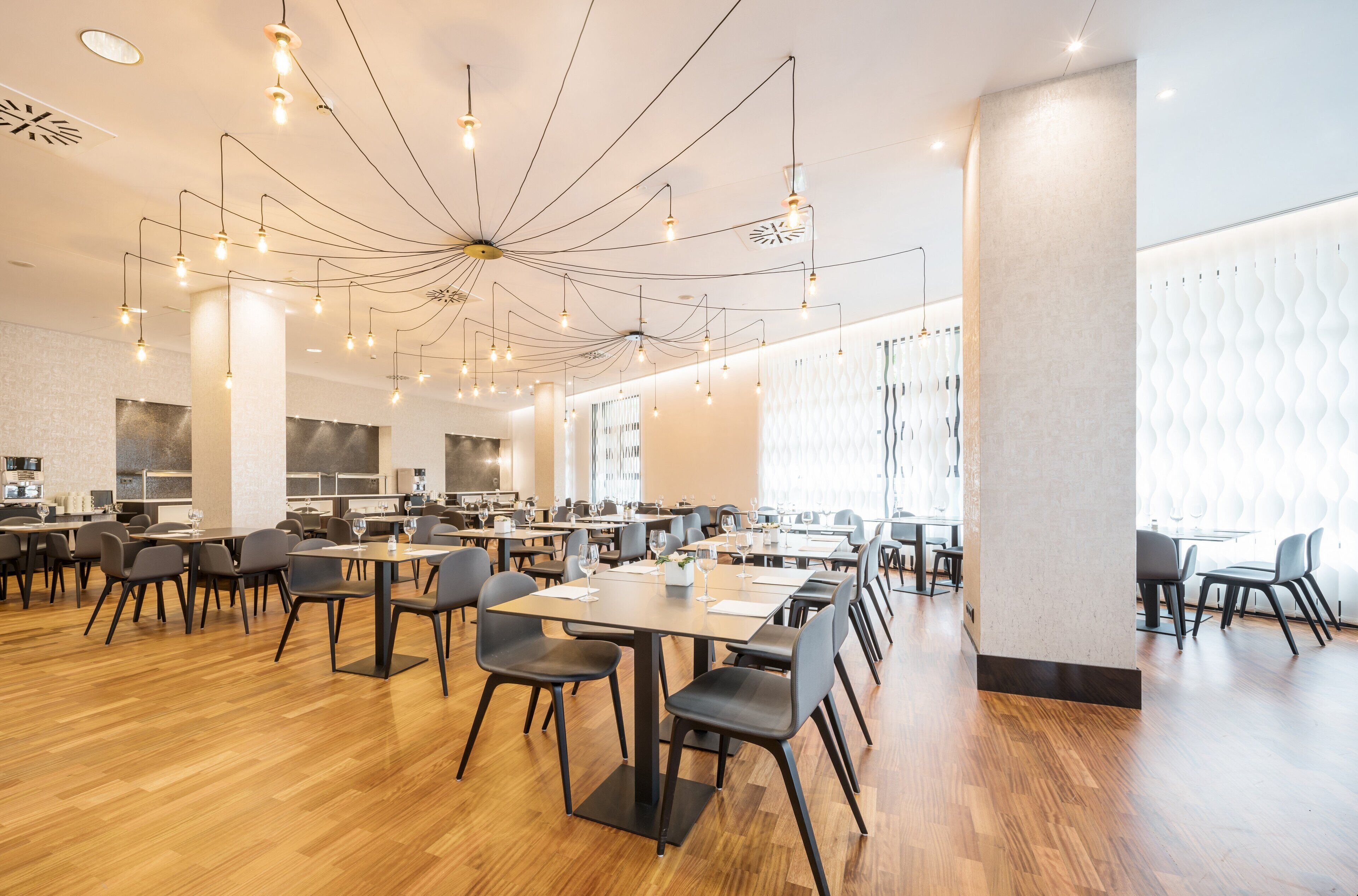 A spacious dining area with multiple tables and chairs, a modern ceiling design with hanging lights, and large windows providing natural light.