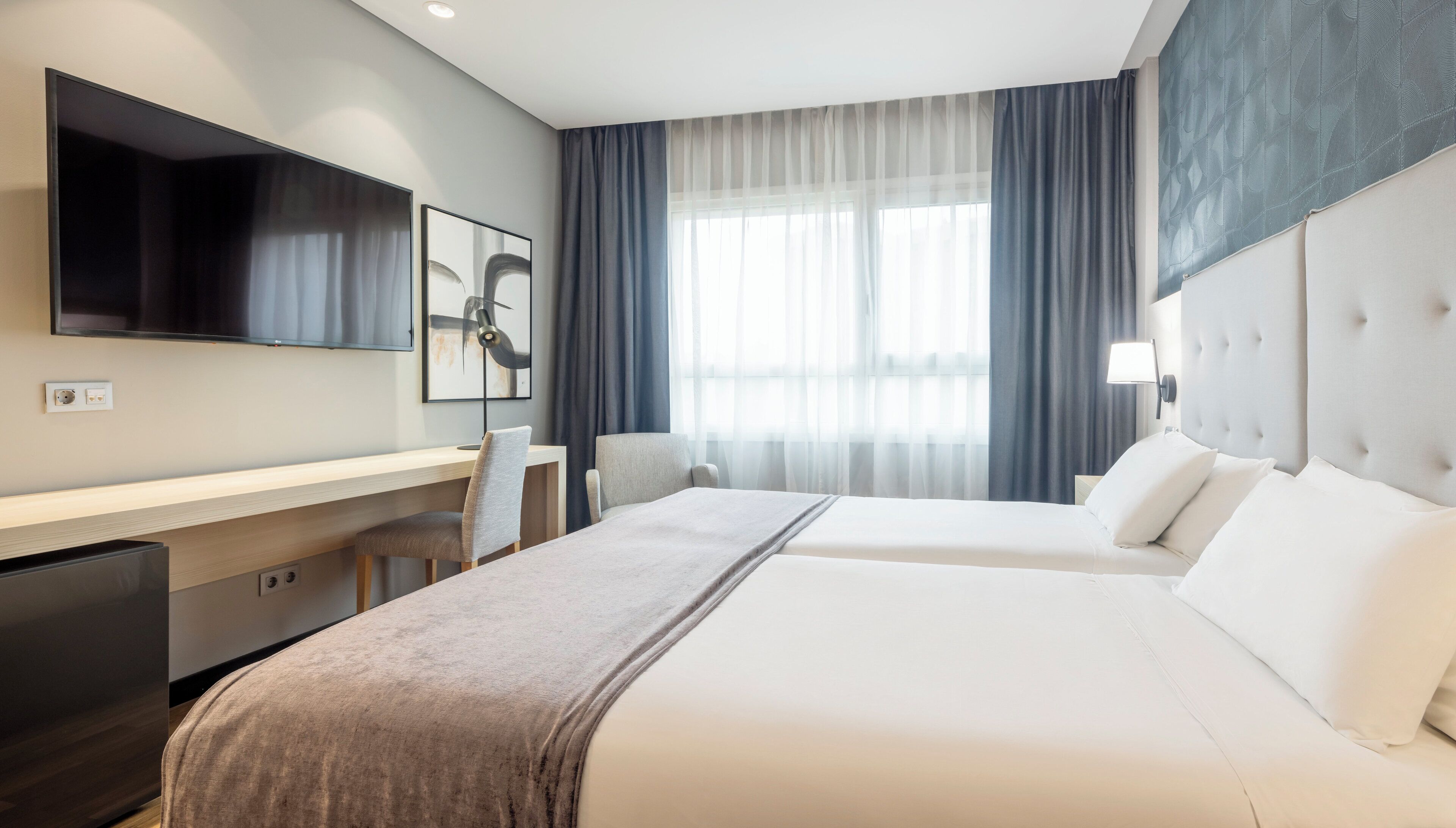 Une chambre d’hôtel avec un lit, un bureau, une chaise, une télévision et une fenêtre avec des rideaux.