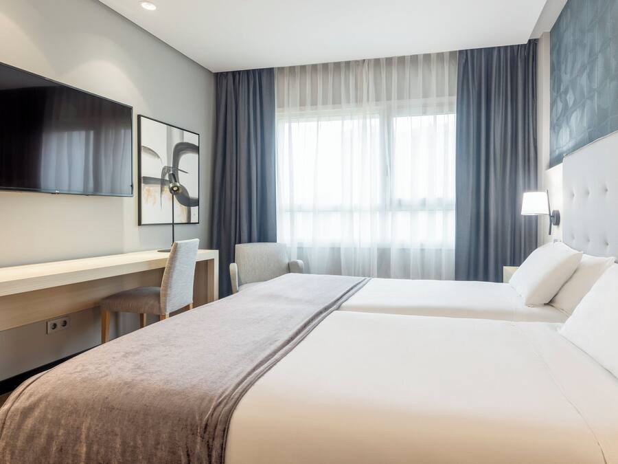 Habitación de hotel con cama, escritorio, silla, televisión y ventana con cortinas.