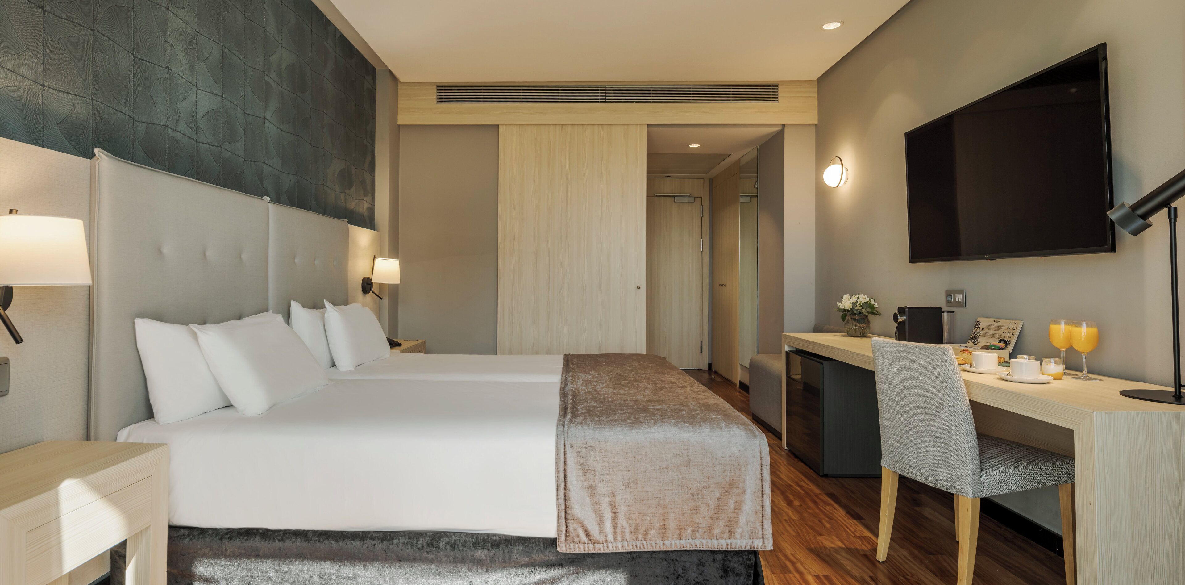 A hotel room with a bed, bedside tables, a desk, a chair, a TV, and a coffee machine.