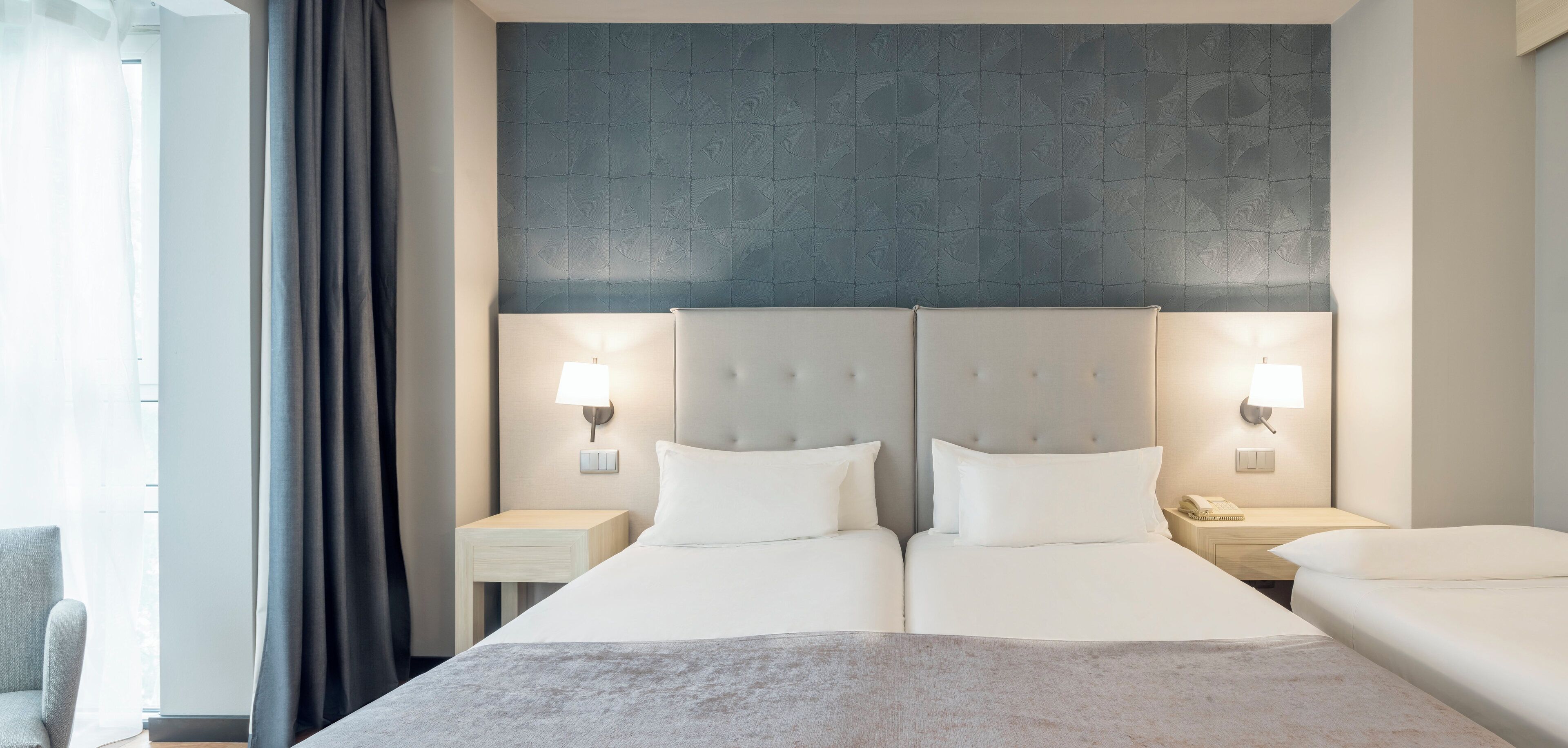 Una habitación de hotel con dos camas, un ventanal con cortinas y una pared texturizada detrás de las camas.