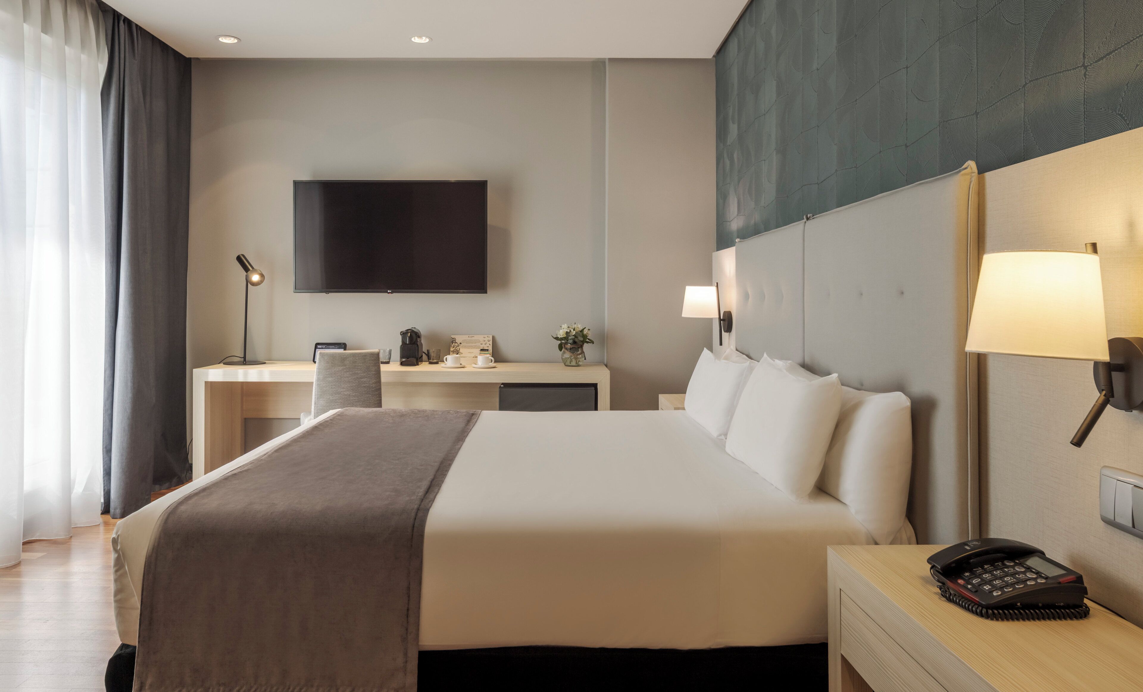 Una habitación de hotel con cama, mesita de noche, escritorio, silla, lámpara y televisor de pantalla plana en la pared.