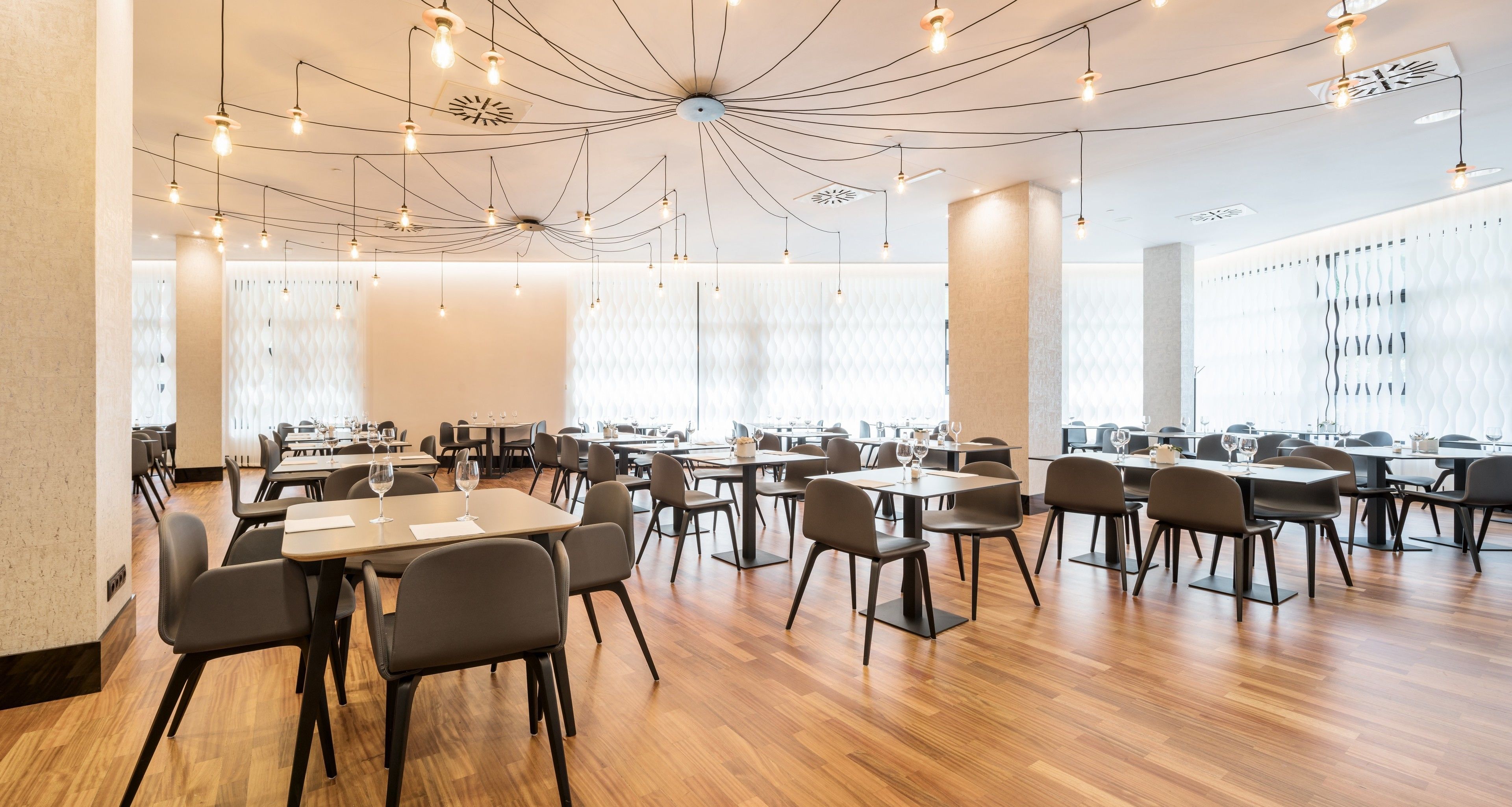 Un comedor espacioso con varias mesas y sillas, lámparas modernas y grandes ventanales que dejan entrar luz natural.