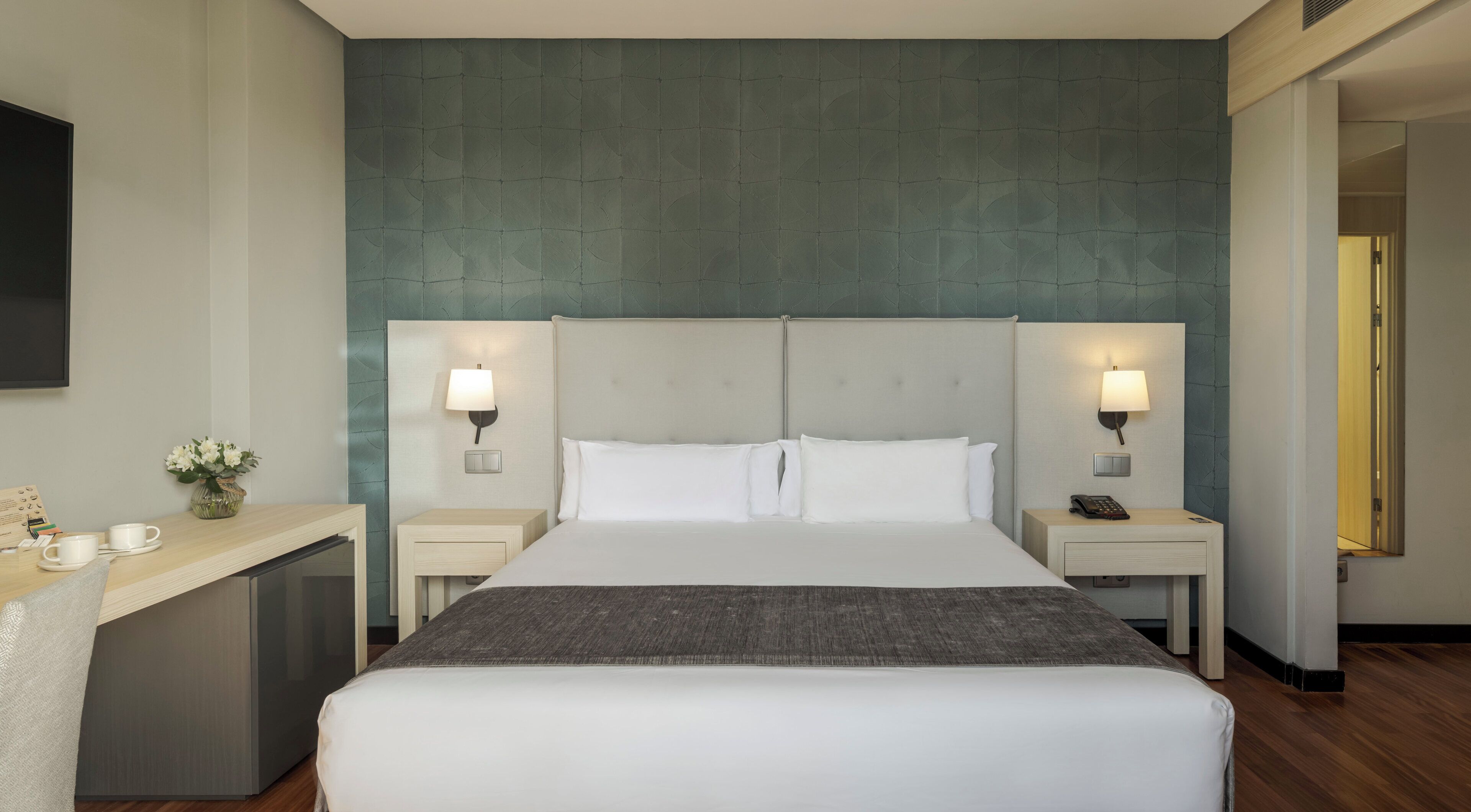 Habitación de hotel con una cama grande, dos mesitas de noche, un escritorio y una lámpara de pared.