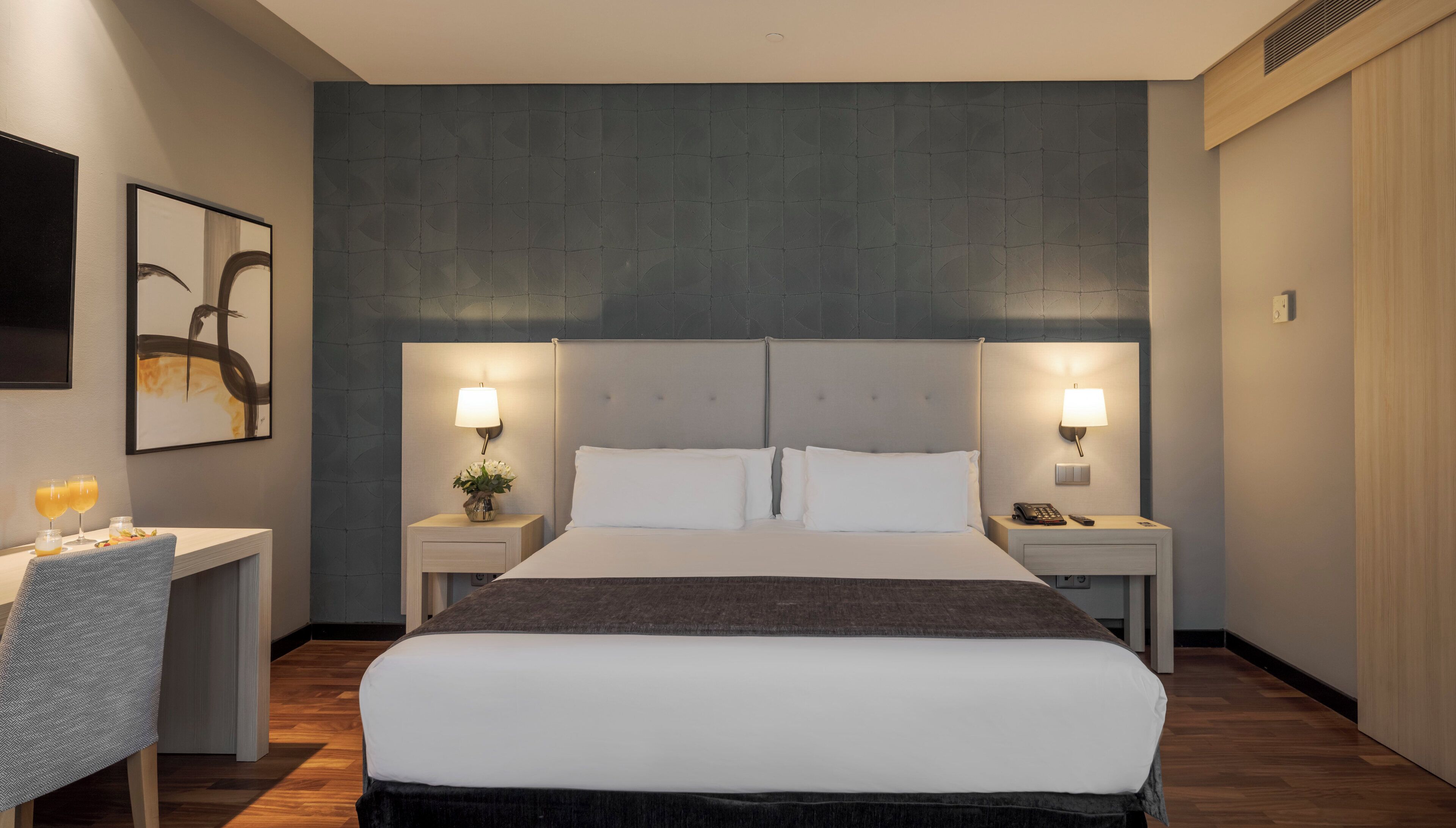 A hotel room with a large bed, two bedside tables, a desk with a chair, and a wall-mounted lamp.