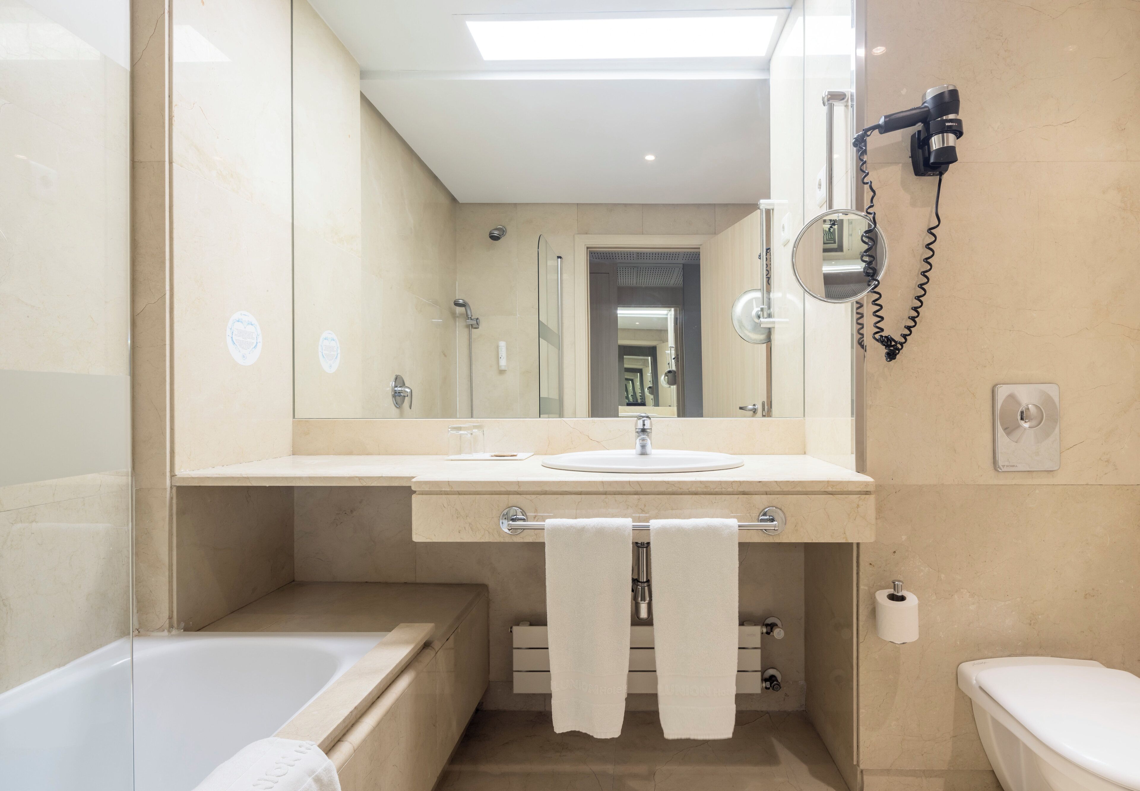Un baño moderno con una bañera, un lavamanos y un inodoro. Se aprecia un perchero con dos toallas, un secador de pelo y un espejo.