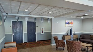 A lobby with a checkered ceiling, two doors, a fire extinguisher, a painting, a sofa, and patterned chairs. - Quality Suites (Indianapolis)