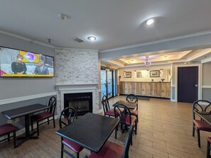 A dining area with a fireplace, a television displaying a news program, and a reception desk with framed pictures. - Quality Suites (Indianapolis)