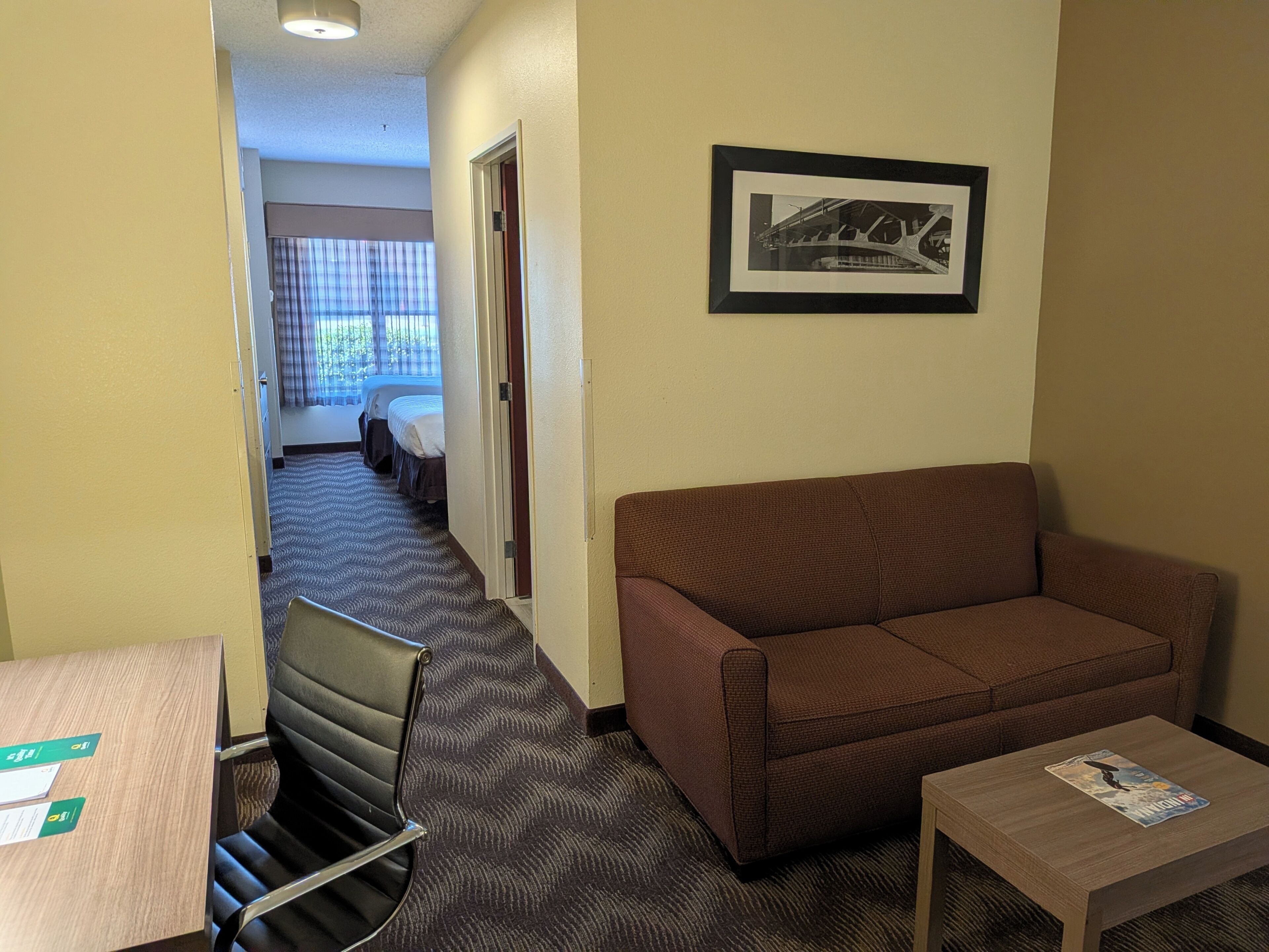 A hotel room with a brown sofa, a small wooden table, a chair, and a framed picture on the wall.
