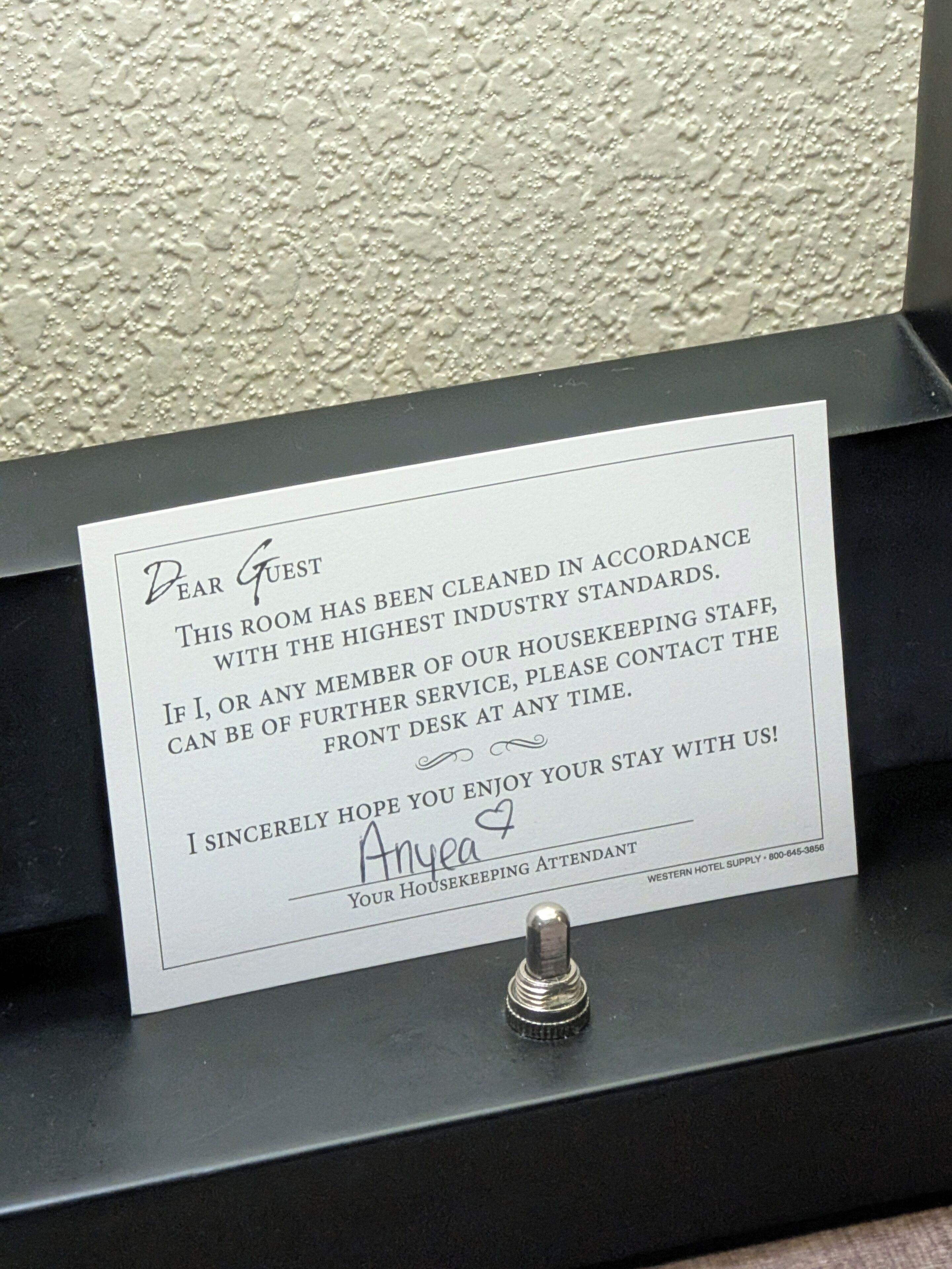 Un petit mot écrit à la main sur une carte, placé dans un porte-carte noir, probablement dans une chambre d’hôtel.
