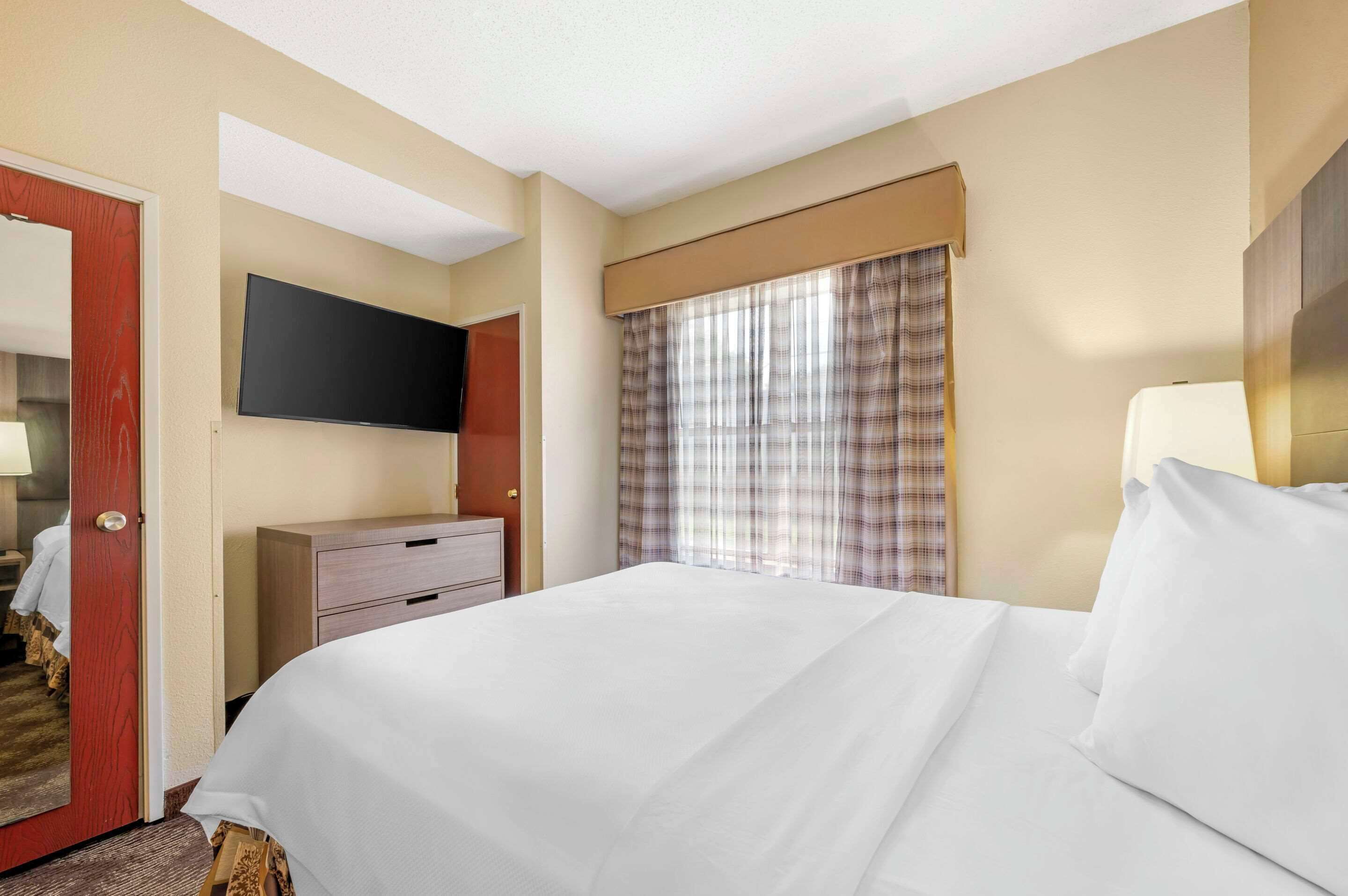 Ein Hotelzimmer mit Bett, Wandfernseher, Kommode und einem Fenster mit Vorhängen.