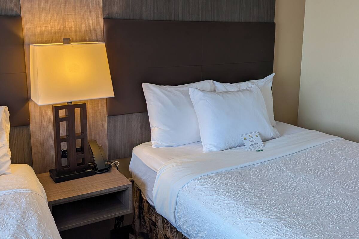 A hotel room with a bed, a bedside table with a lamp, and a small sign on the pillow.