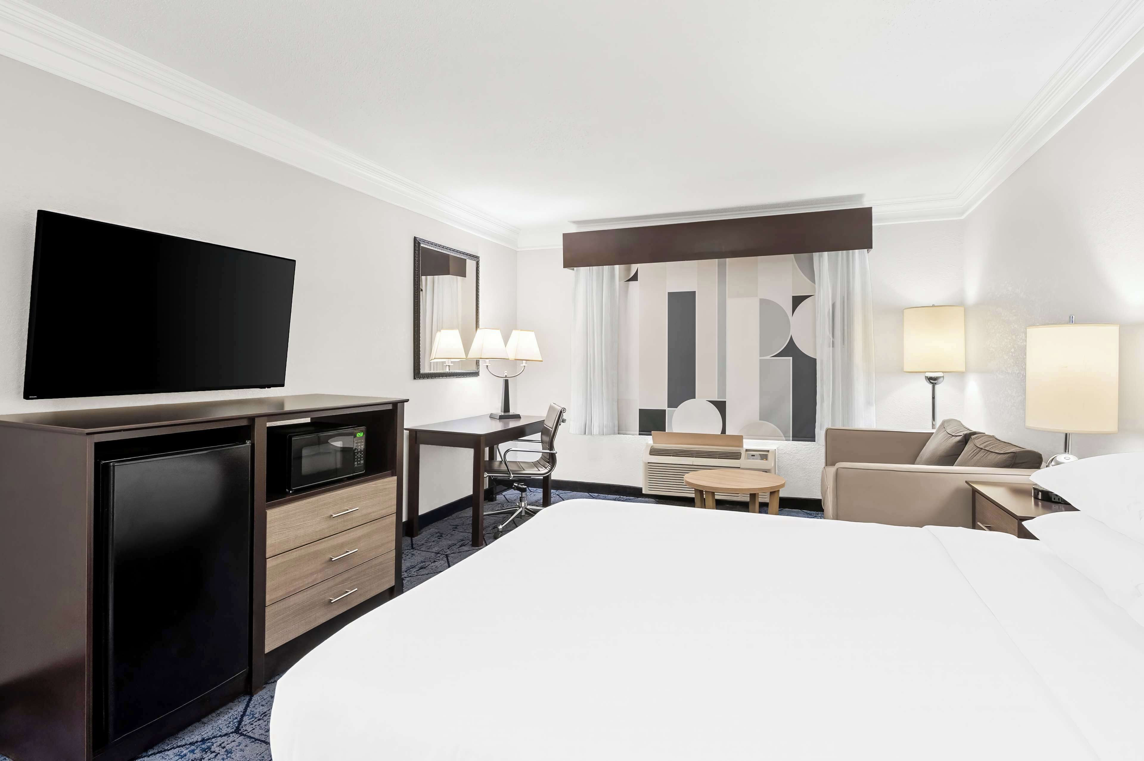 A hotel room with a large bed, a desk with a flat-screen TV, a microwave, a chair, a small table, and two lamps.