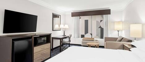 A hotel room with a large bed, a desk with a flat-screen TV, a microwave, a chair, a small table, and two lamps.