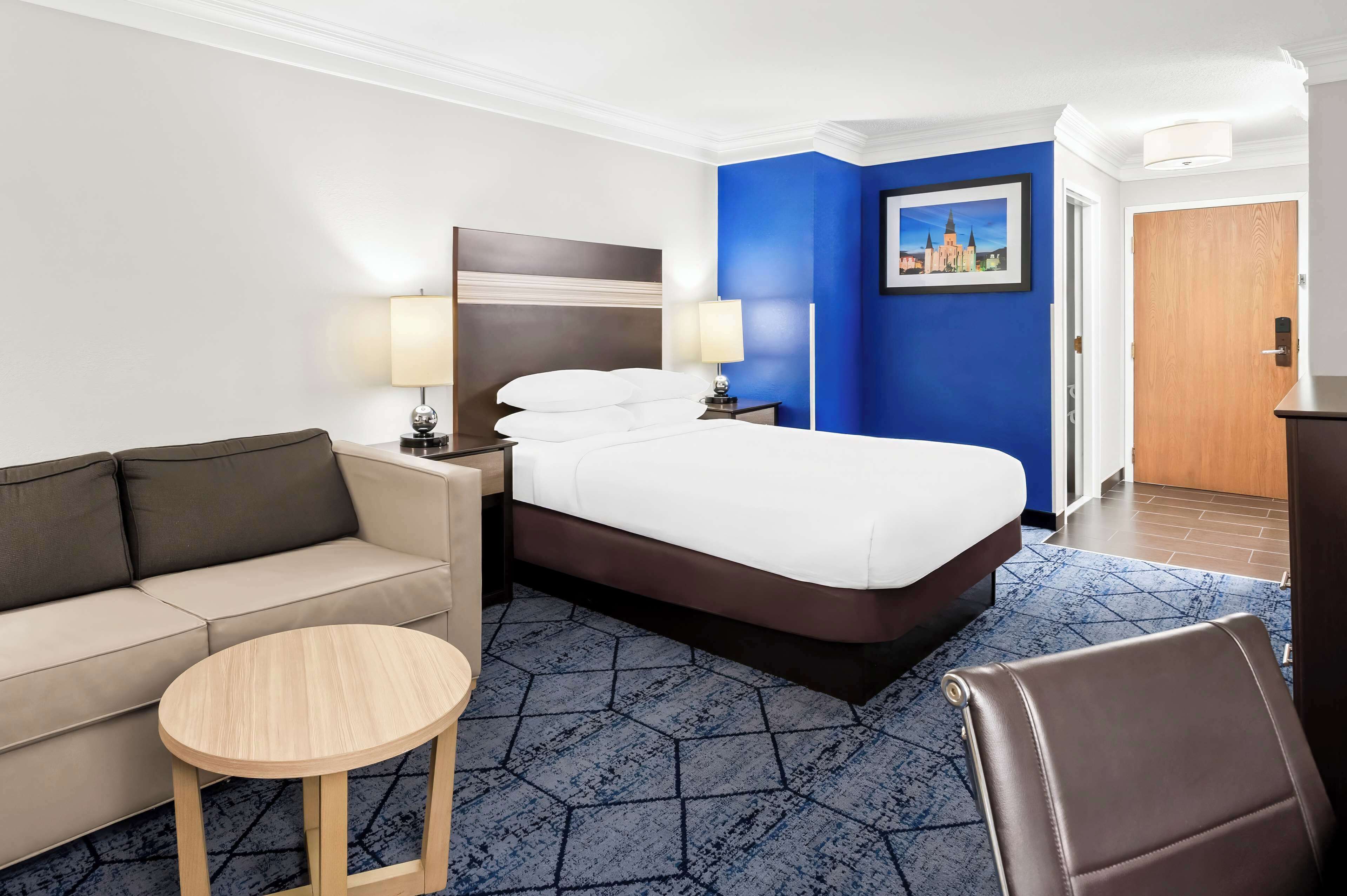 Ein Hotelzimmer mit Bett, Sofa, kleinem runden Tisch, Sessel, Nachttisch mit Lampen und blauer Akzentwand.