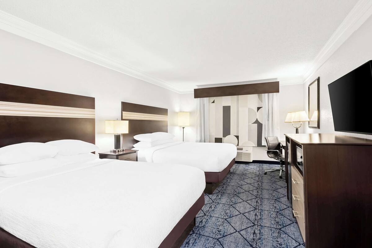A hotel room with two beds, a desk, a chair, a TV, and a large window.
