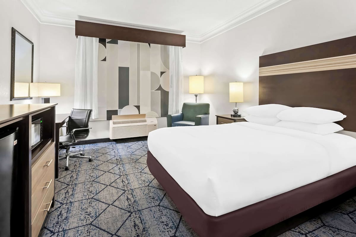 A hotel room with a large bed, a desk with a computer, a chair, a lamp, and a large window with curtains.