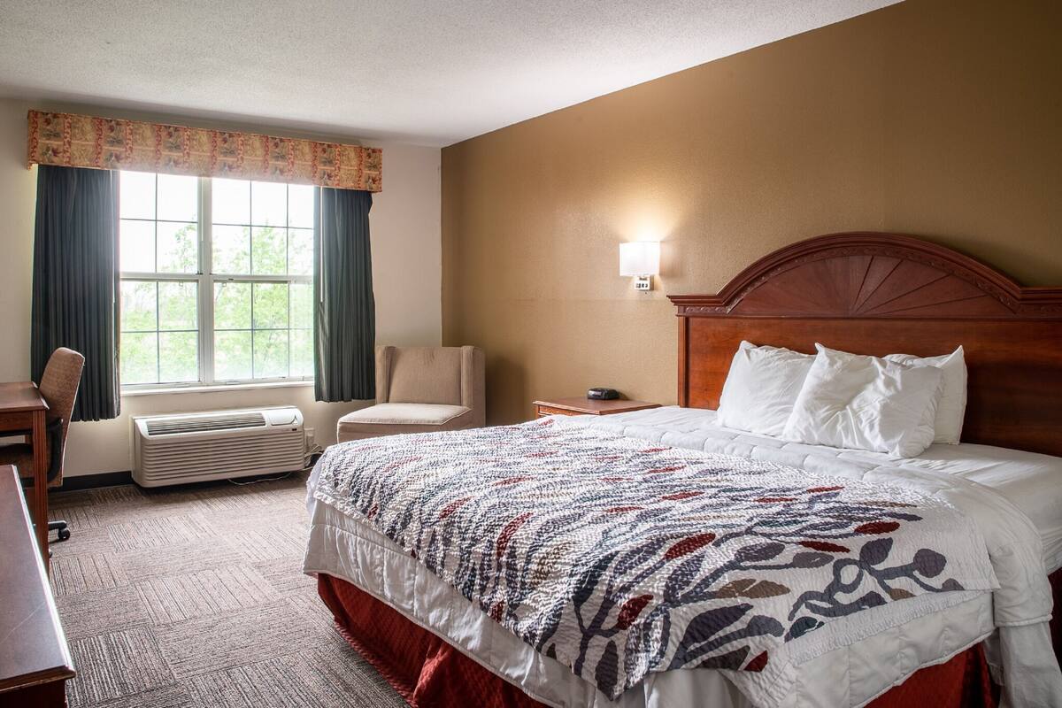 A hotel room with a large bed, a desk, a chair, a window with curtains, and a wall-mounted lamp.