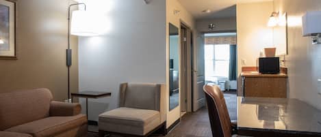 A hotel room with a sofa, a small armchair, a dining table with chairs, a flat-screen TV, and a kitchenette area.