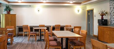 A dining area with wooden tables and chairs, a potted plant, and a kitchen area with a refrigerator and cabinets.