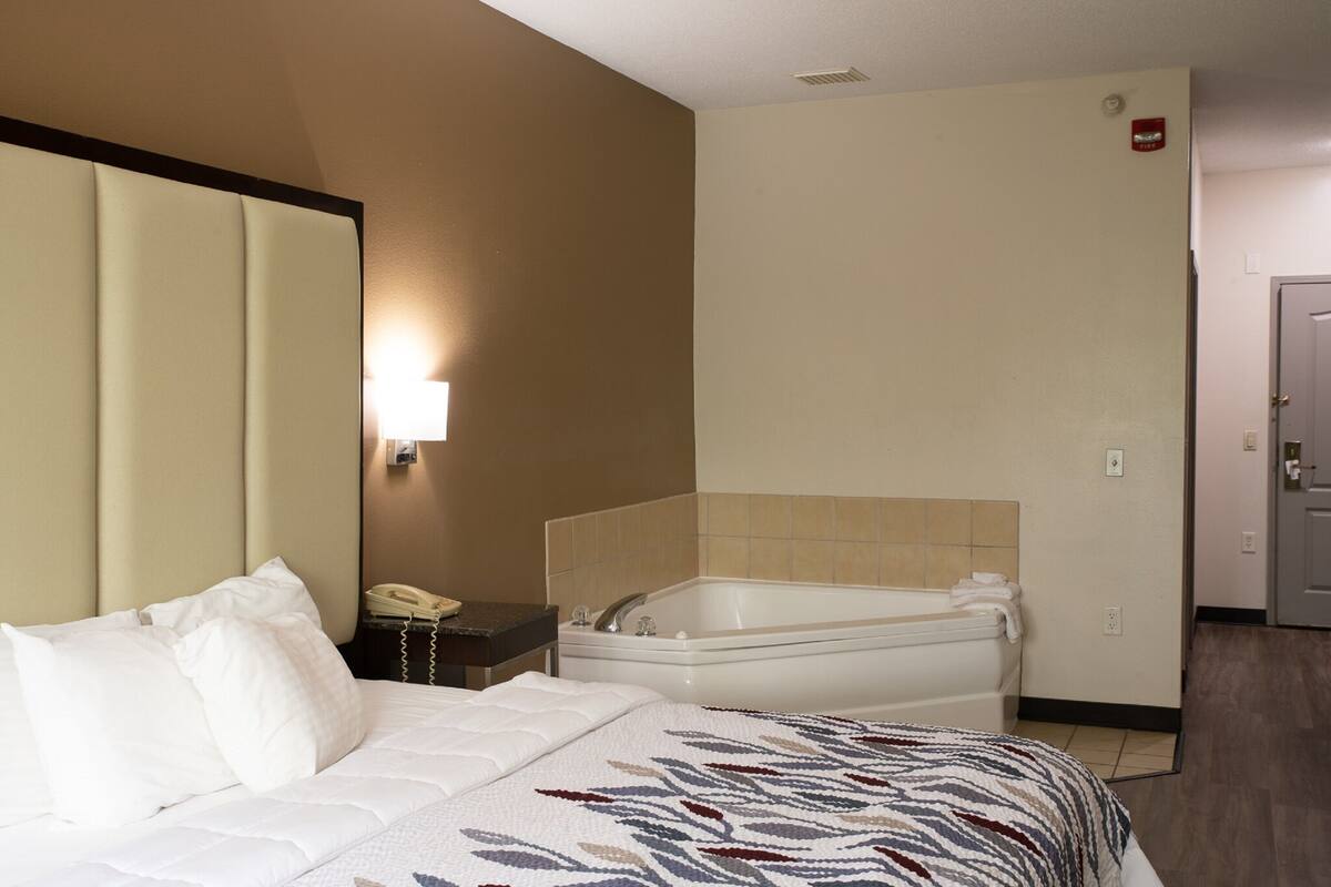 A hotel room with a bed, a bedside table with a telephone, a wall-mounted light, and a bathtub.