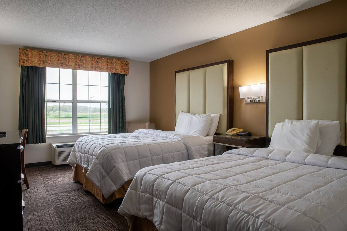 A hotel room with two beds, a desk, a chair, a window with curtains, and a nightstand with a lamp.